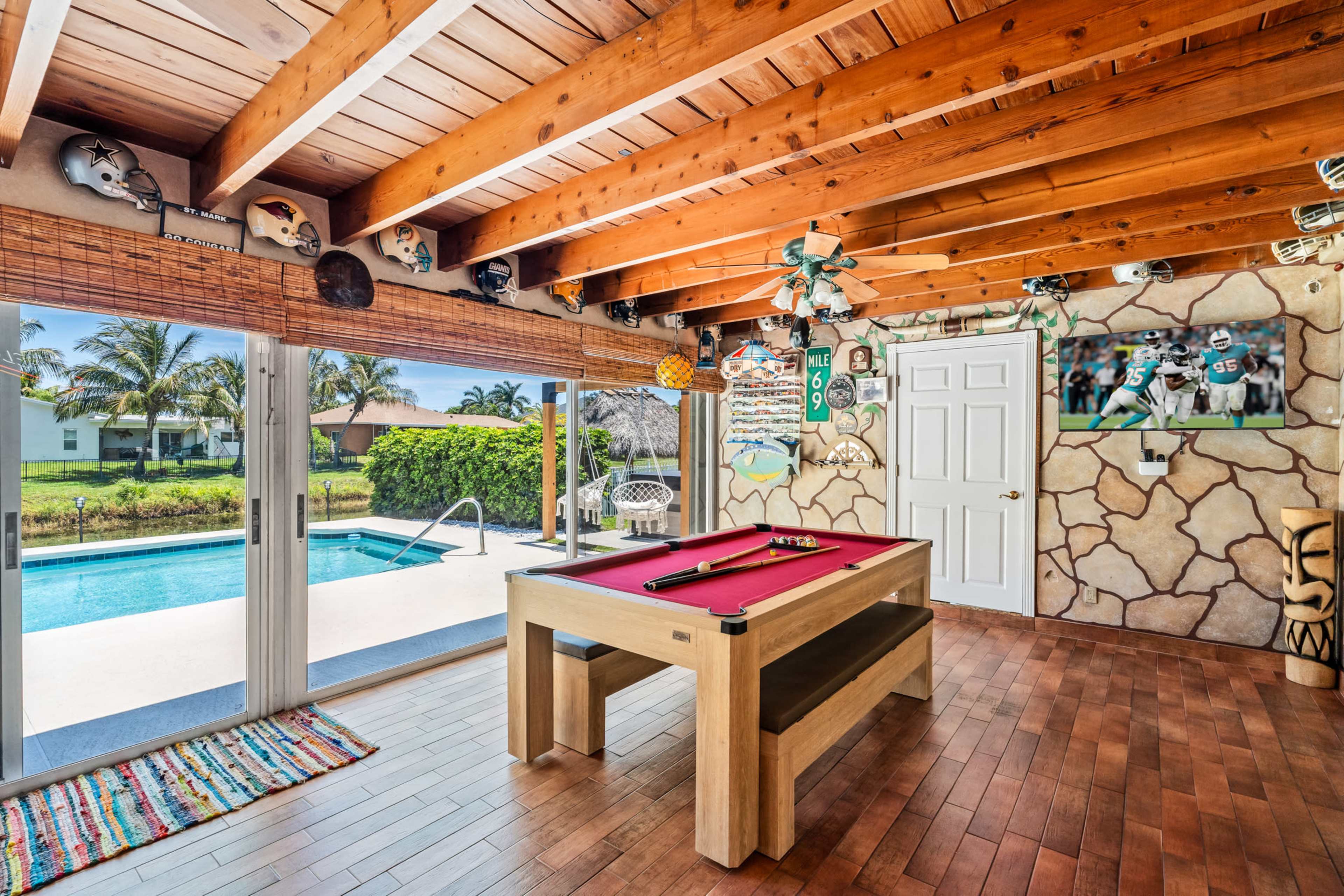 A game room features a pool table with wooden beams overhead and sliding glass doors opening to a pool area surrounded by palm trees.