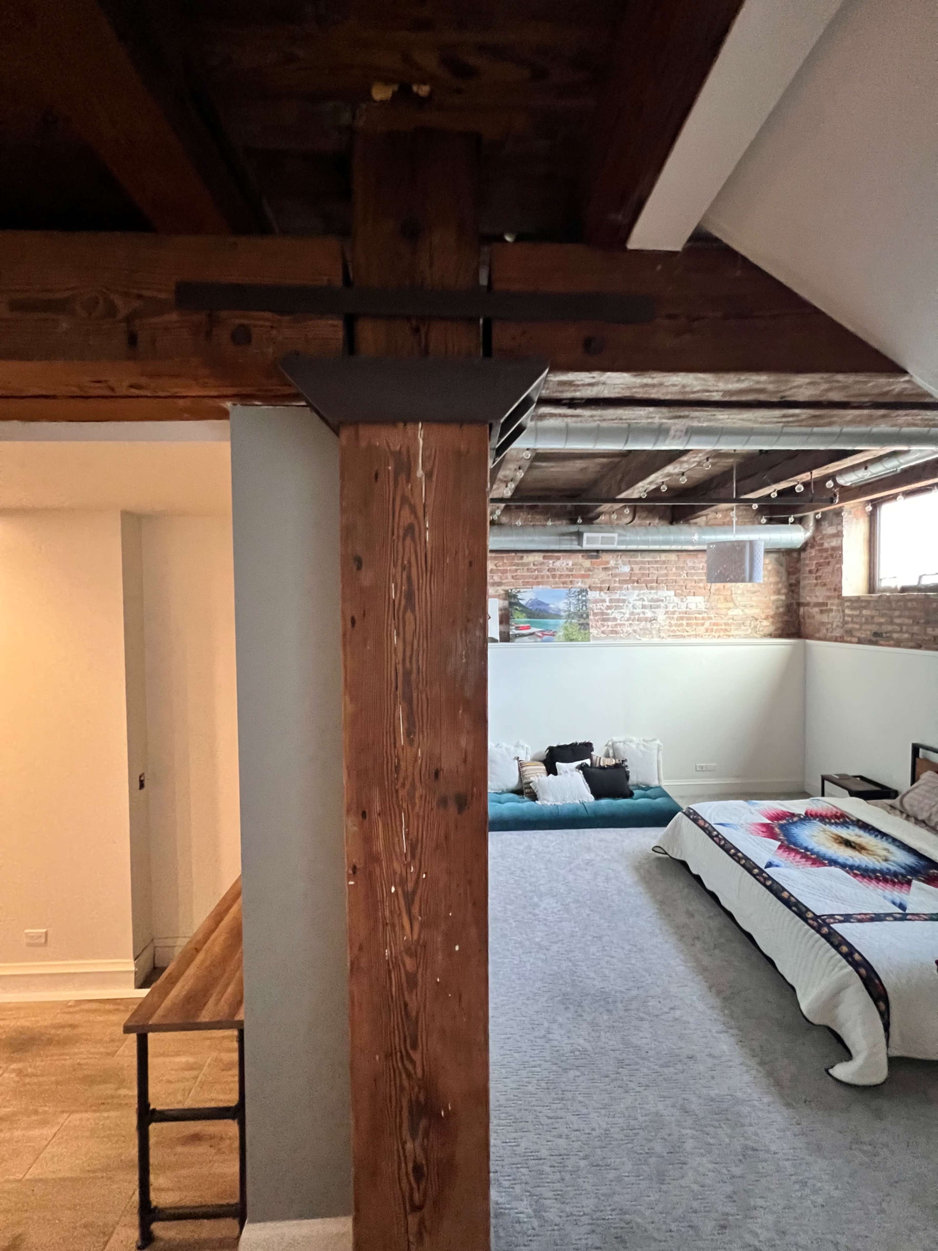 The image shows a room with exposed wooden beams and a spacious layout featuring a bed, decorative items, and a partially visible hallway.
