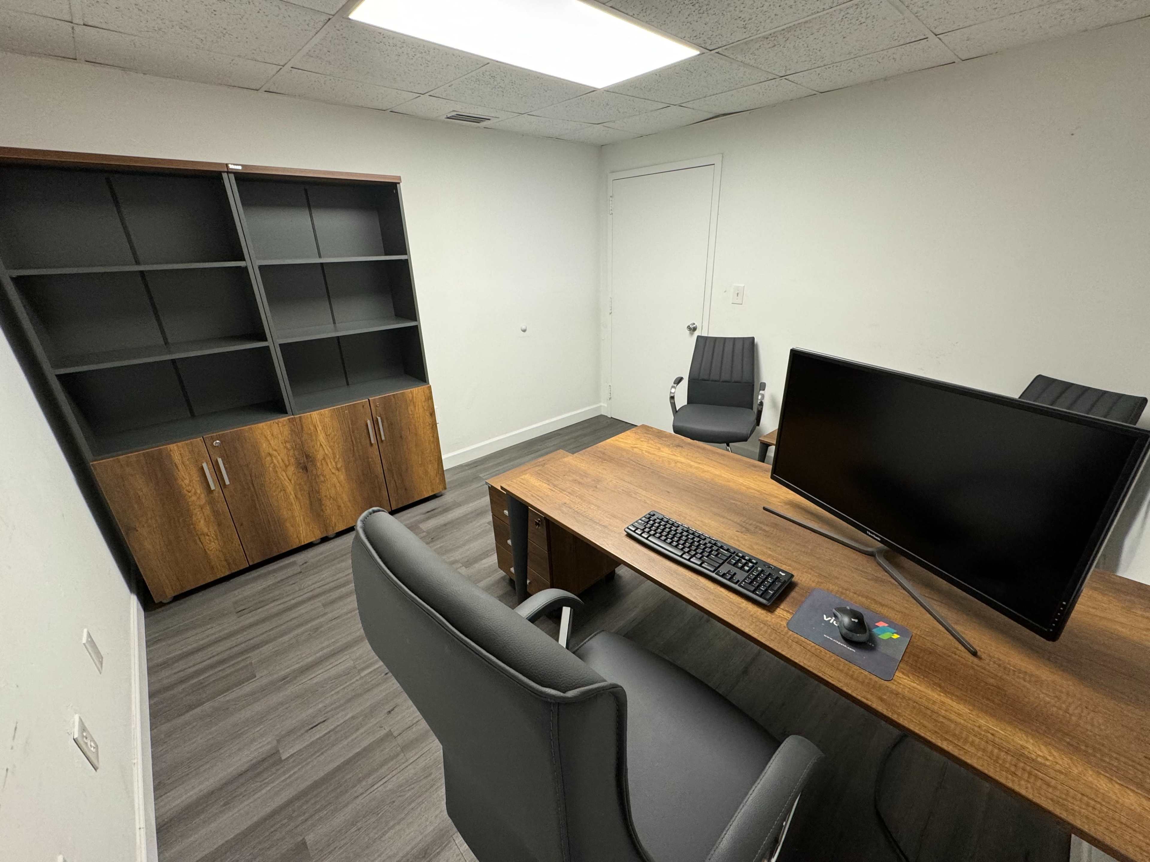 An office space features a wooden desk with a computer and keyboard, two black chairs, and a shelving unit against the wall.