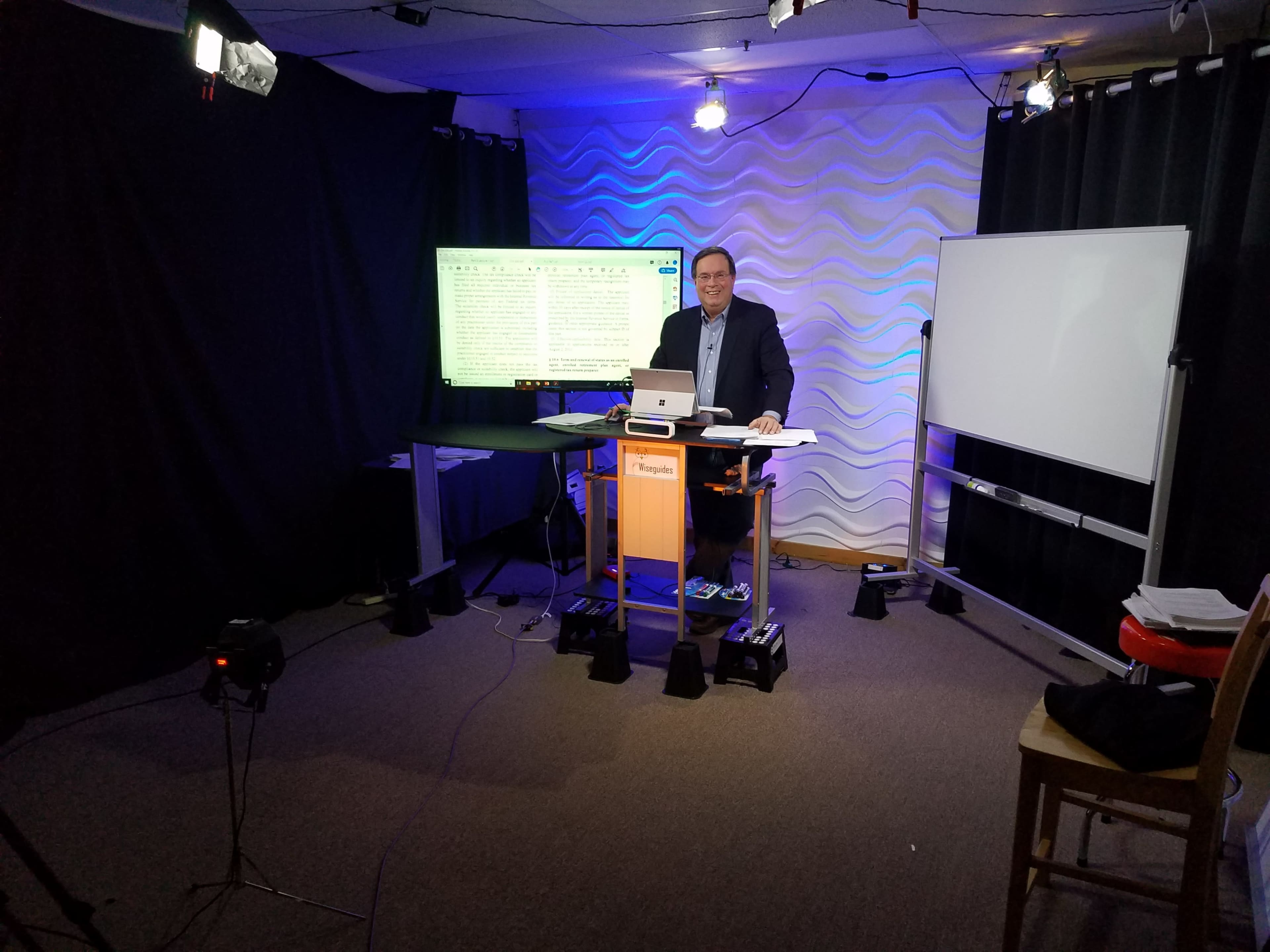 A man stands at a desk with a laptop in a studio setup, surrounded by video equipment and a large screen displaying text.