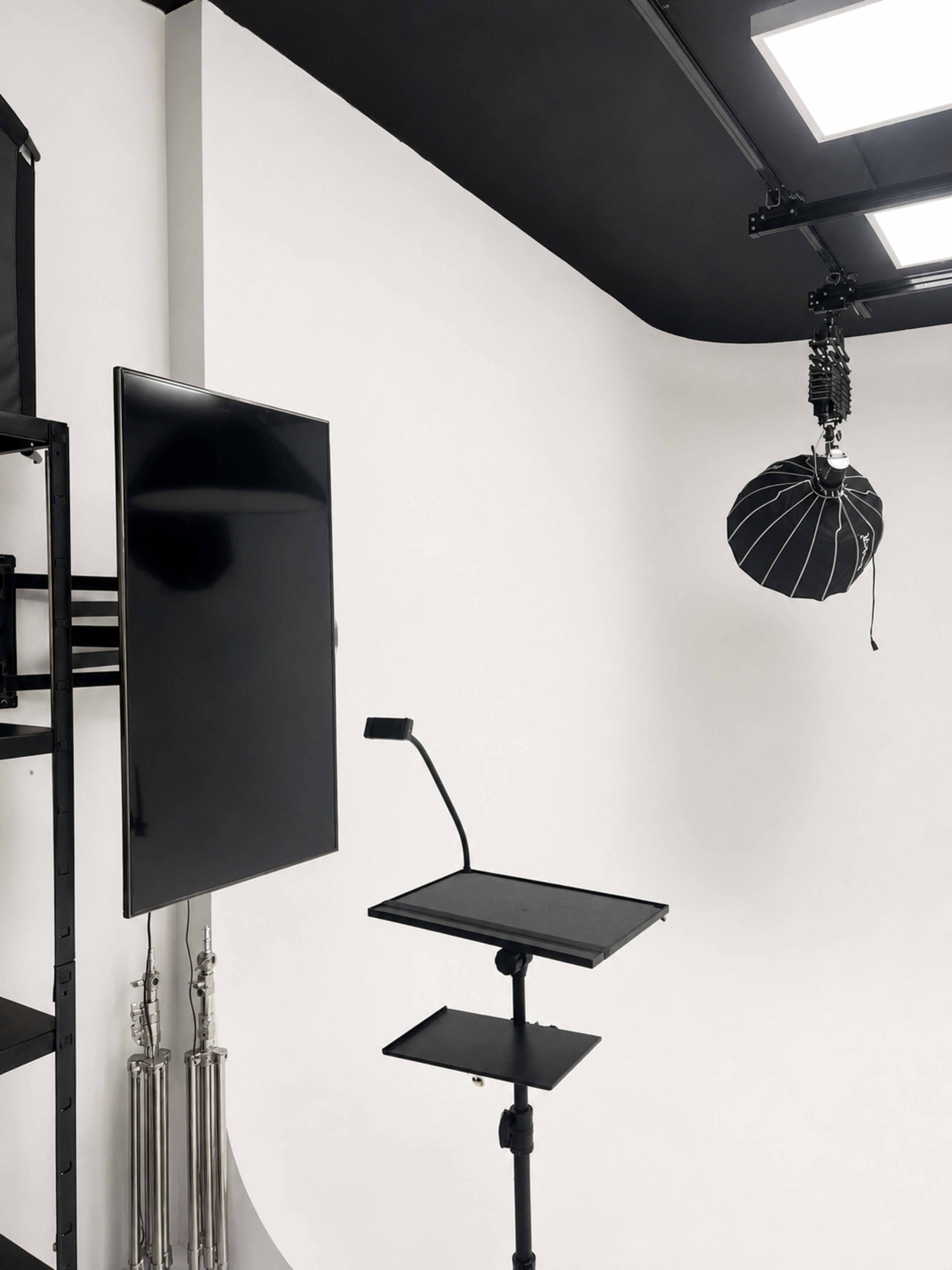 The image shows a photography studio setup with a black monitor, a multi-tiered stand, and a lighting umbrella hanging from the ceiling against a white backdrop.