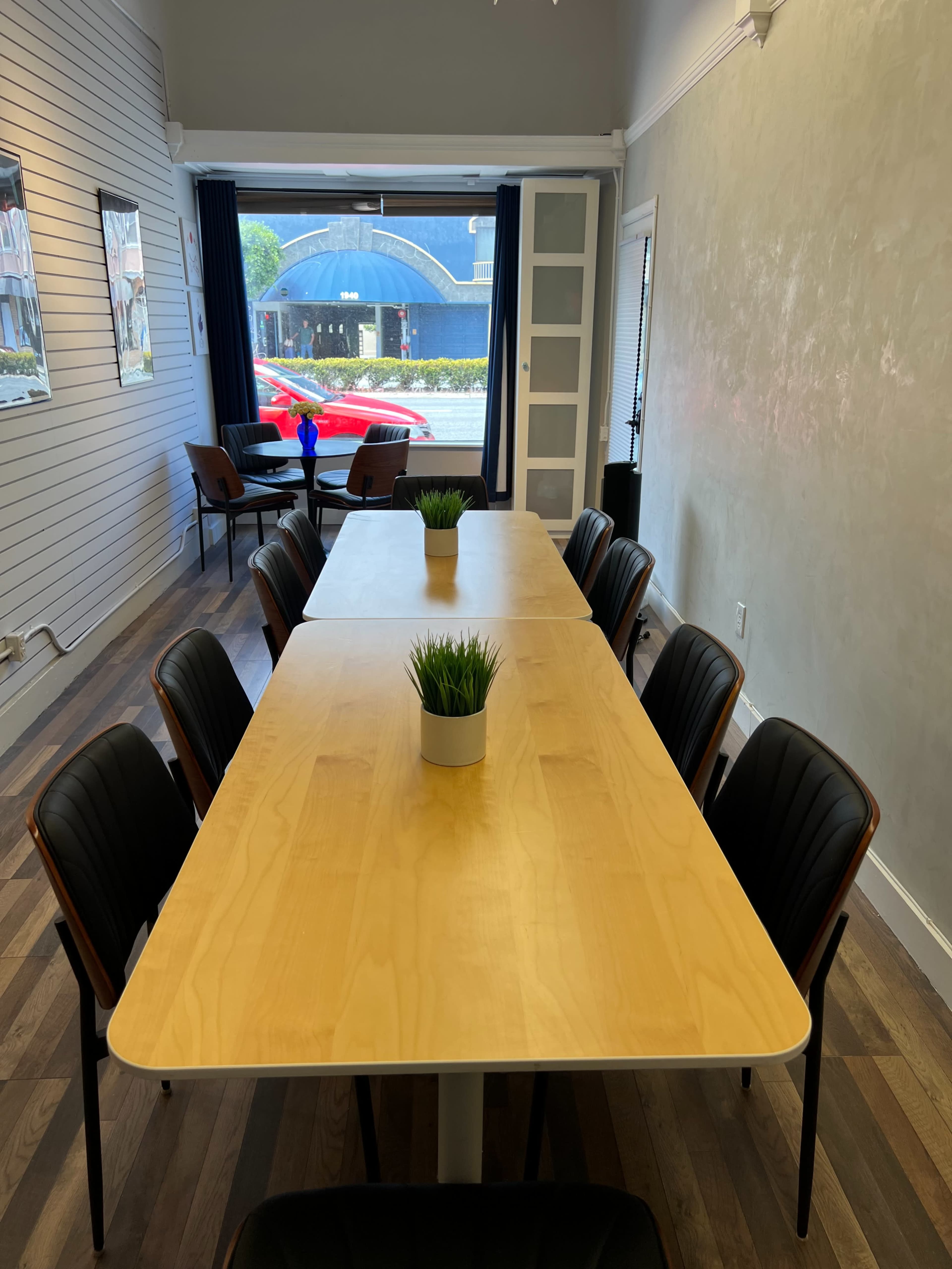 The image shows a well-lit meeting room with a long table surrounded by chairs, and a view of a street through large windows.