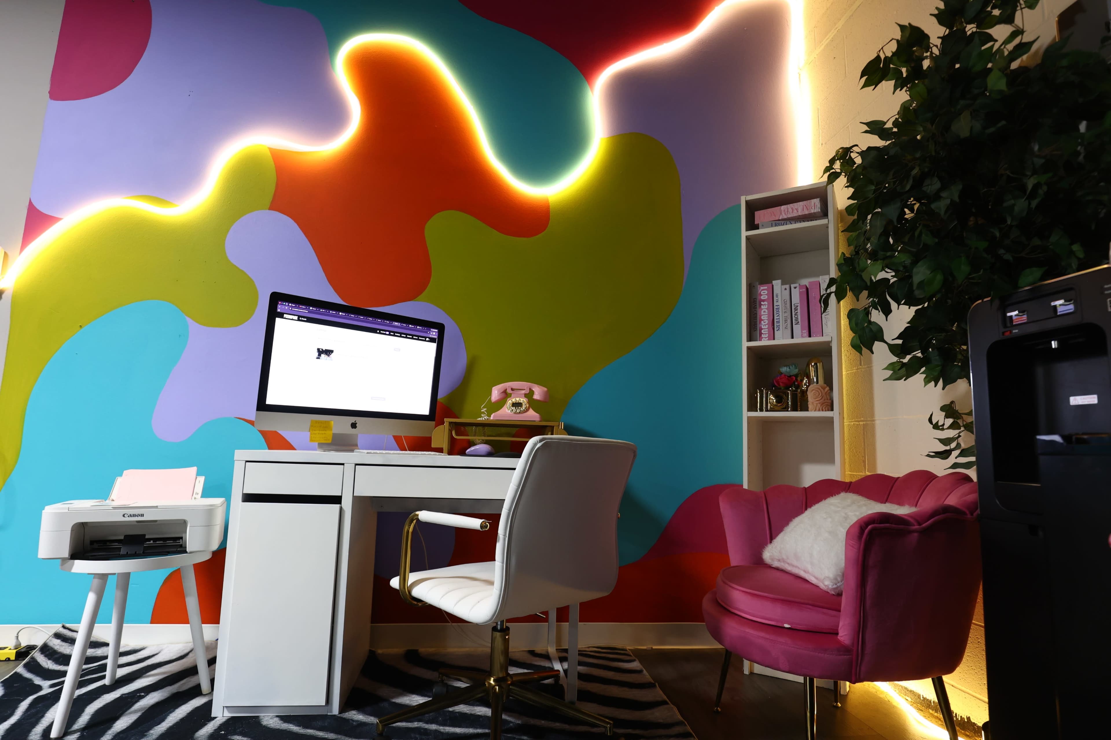 A colorful room features a white desk with a computer, a pink armchair, and a shelf filled with books, against a vibrant abstract mural.