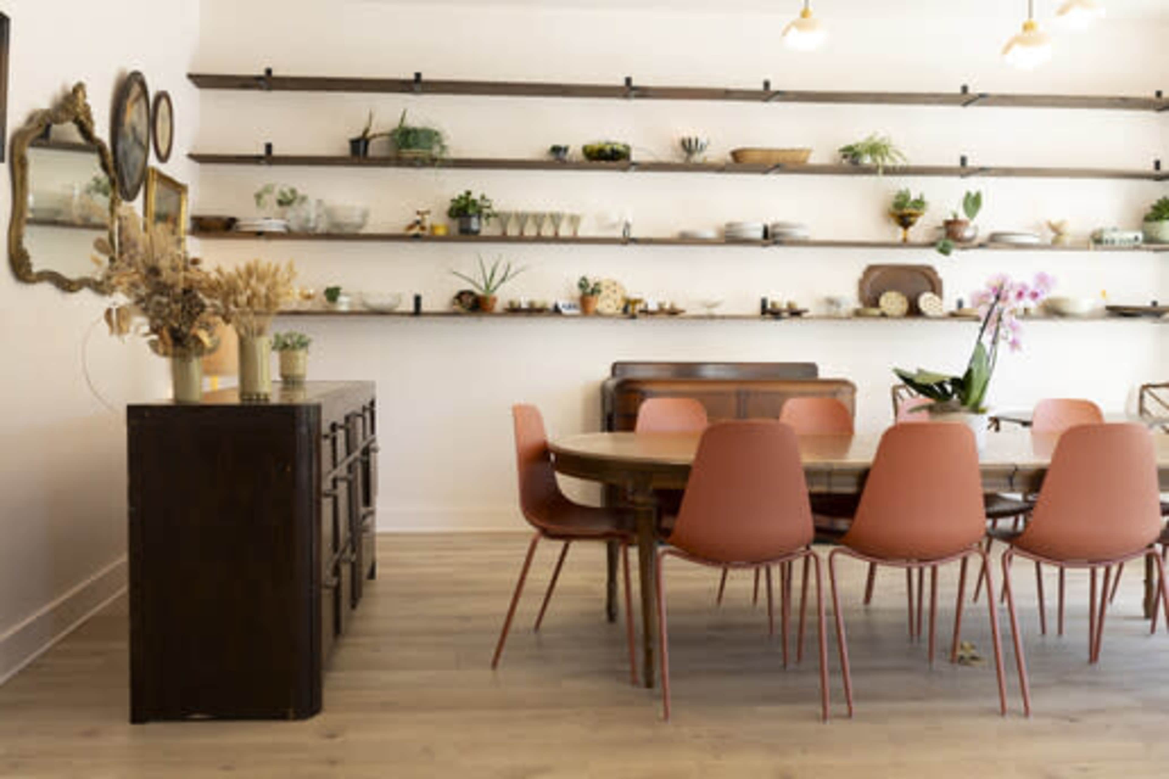 A large wooden dining table surrounded by pink chairs is set against a wall of shelves displaying various decorative items and plants.