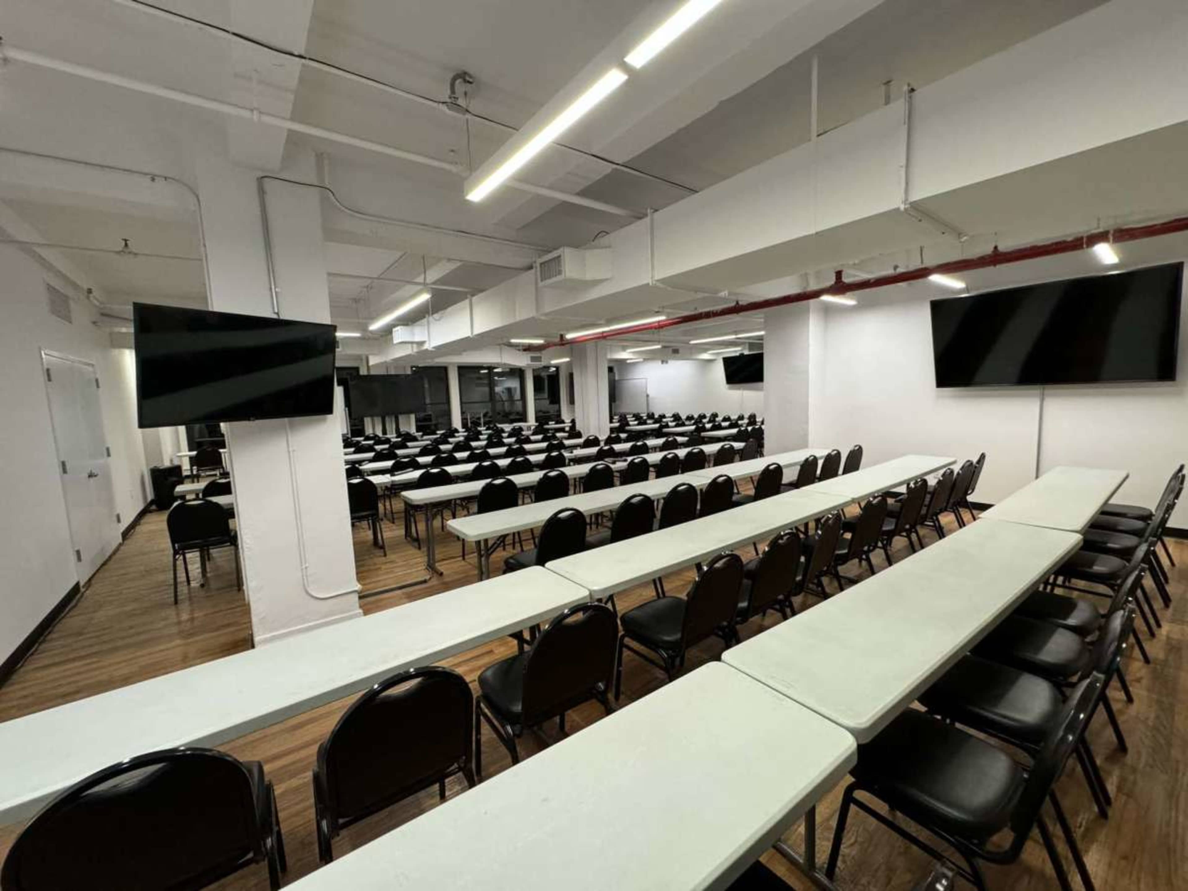 The image shows a spacious classroom or conference area arranged with rows of tables and chairs, equipped with wall-mounted screens.