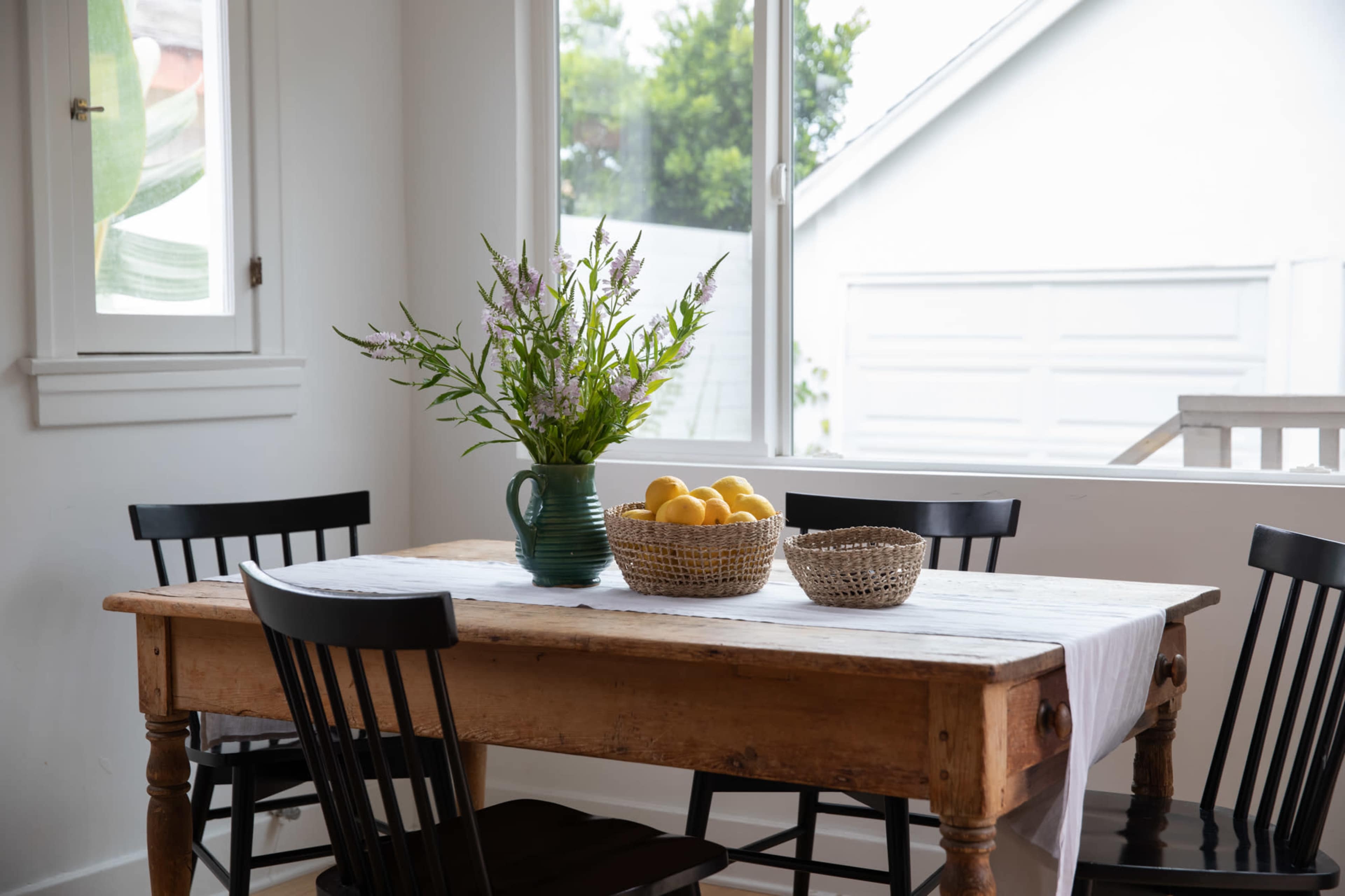 A wooden dining table with a vase of flowers and a bowl of lemons sits surrounded by black chairs in a bright room with large windows.