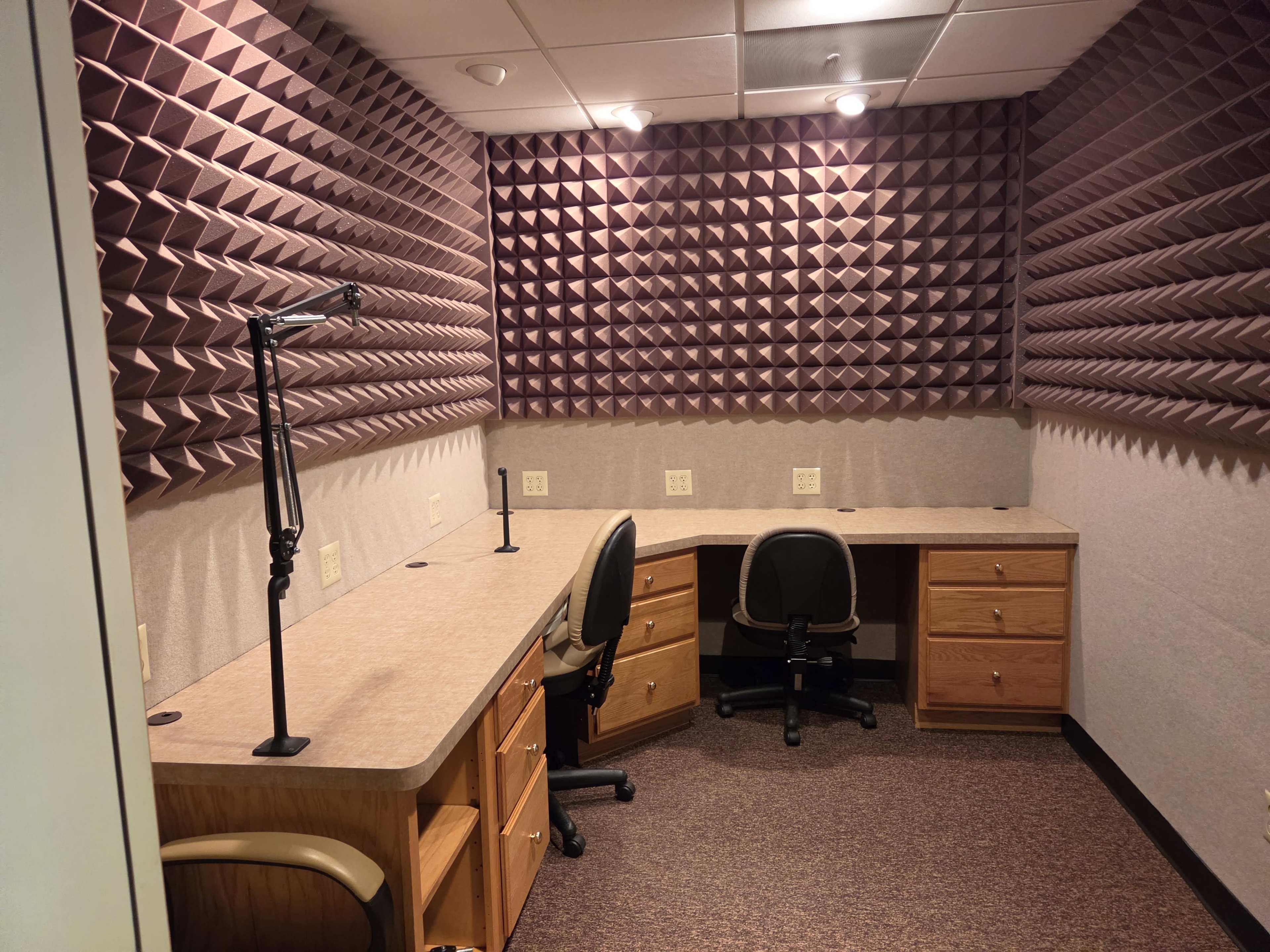 The image shows a soundproof room featuring two desks with chairs, surrounded by acoustic foam wall panels.
