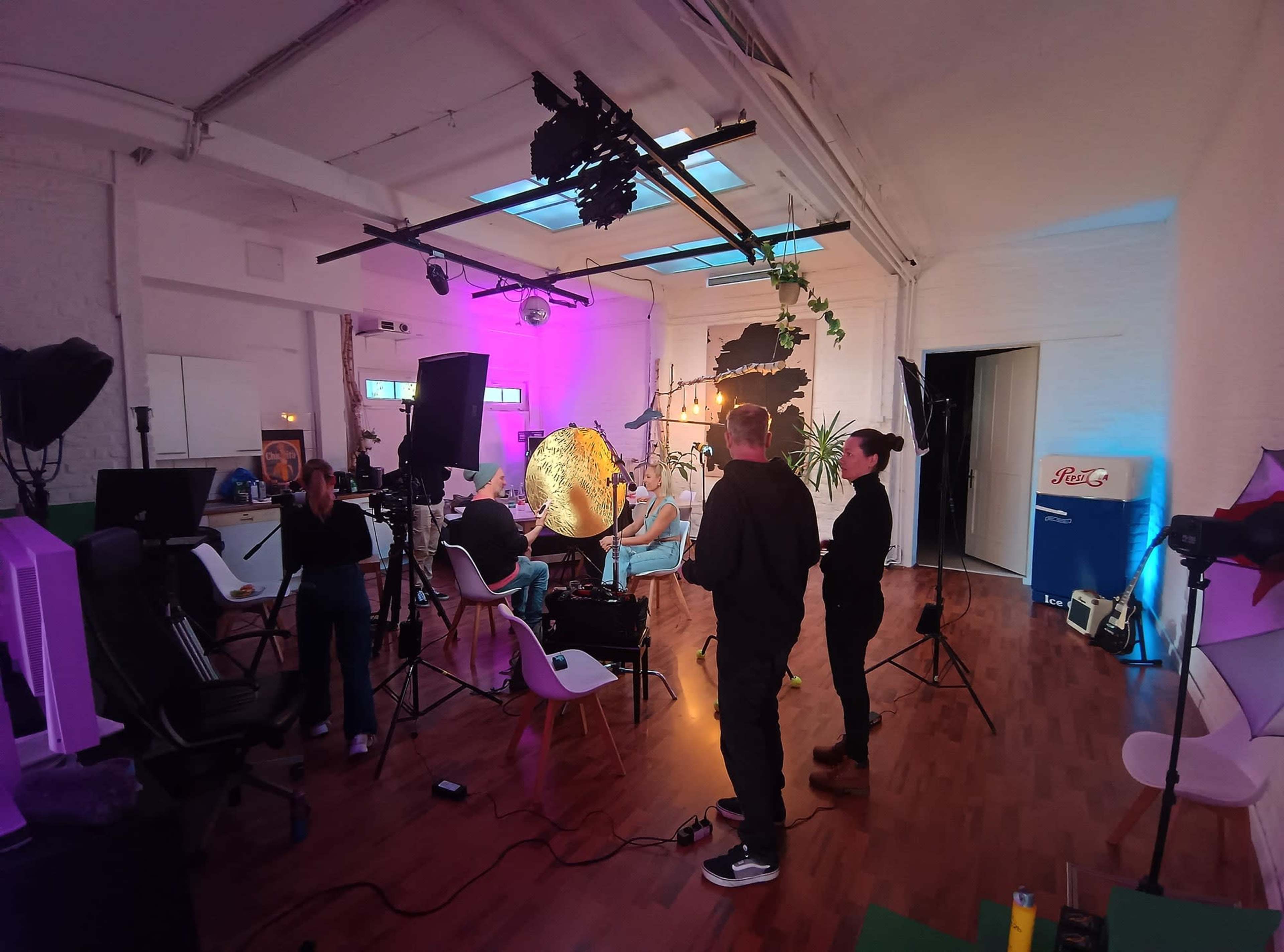 A photography studio is set up with colorful lighting, cameras, and various equipment, while two individuals discuss in the foreground.