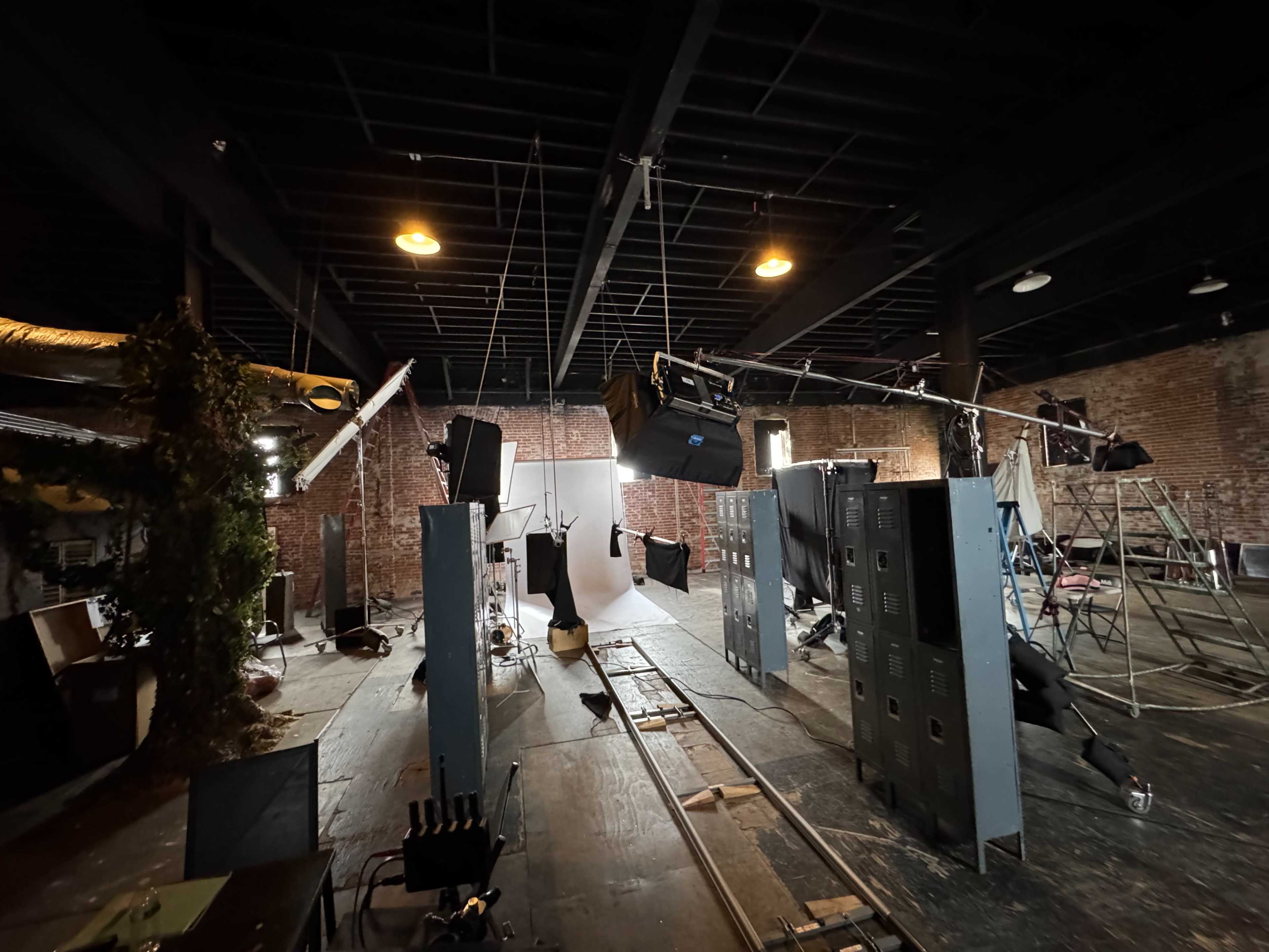 The image shows a spacious studio filled with various equipment, including lighting rigs and backdrops, amidst visible construction elements like brick walls and wooden flooring.