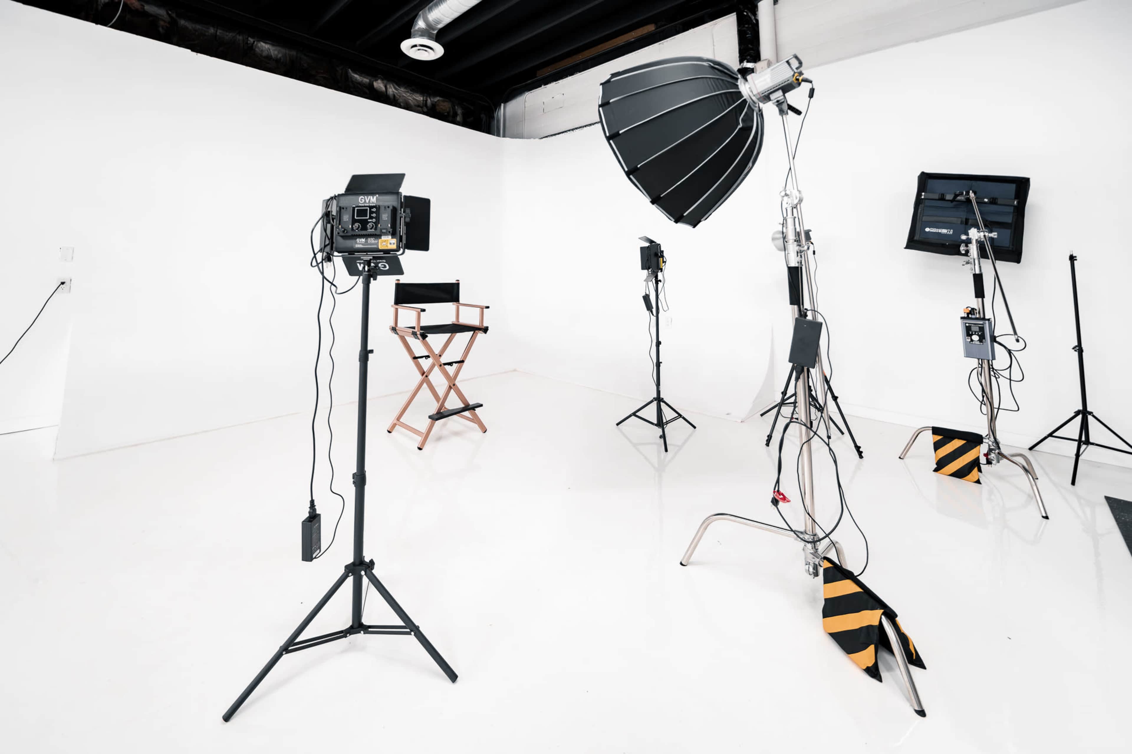 The image shows a photography studio with various lighting equipment, a director's chair, and a clean white backdrop.