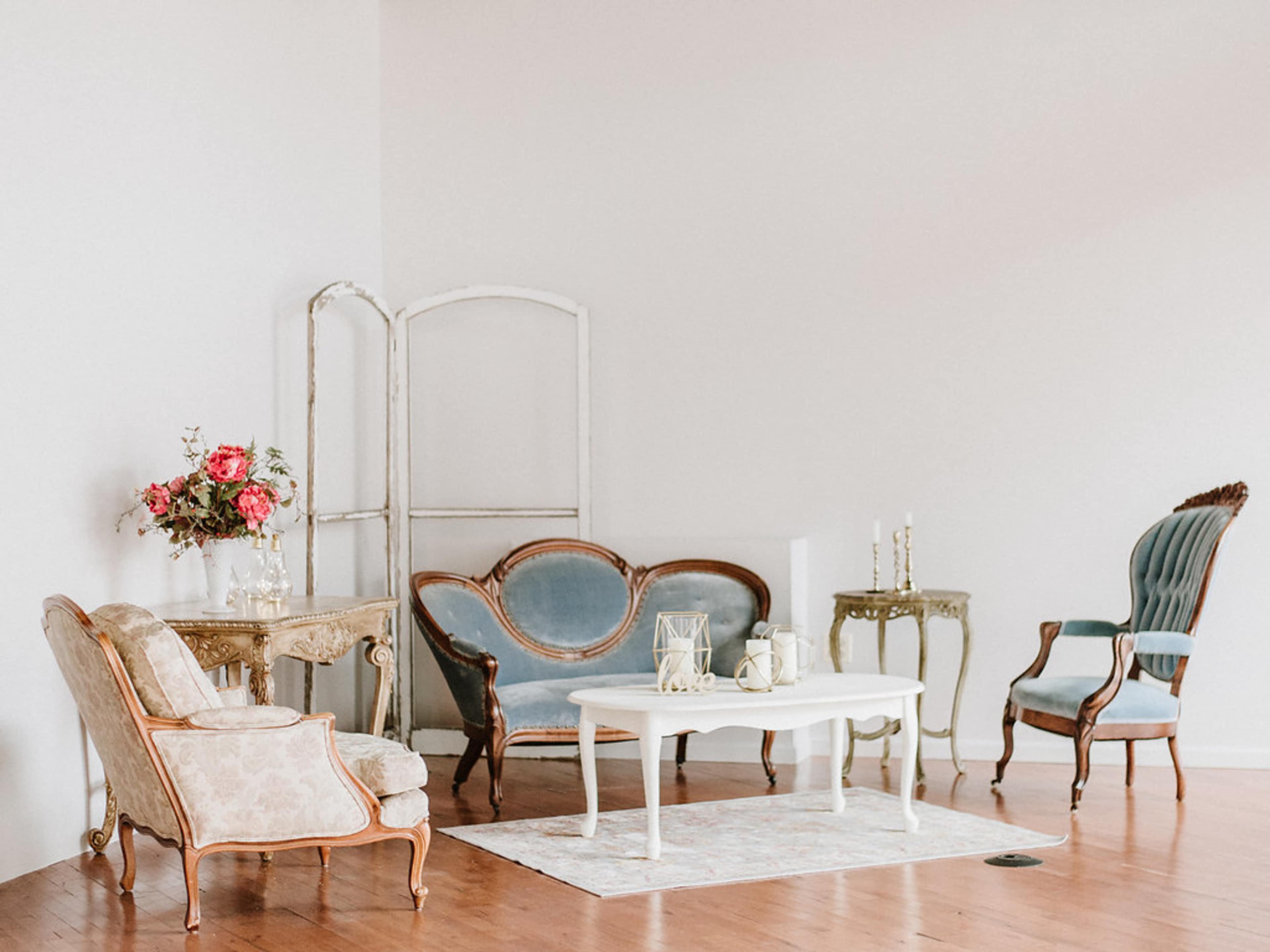 A softly lit room featuring vintage furniture, including a floral sofa, an upholstered chair, and a white coffee table, arranged on a wooden floor.