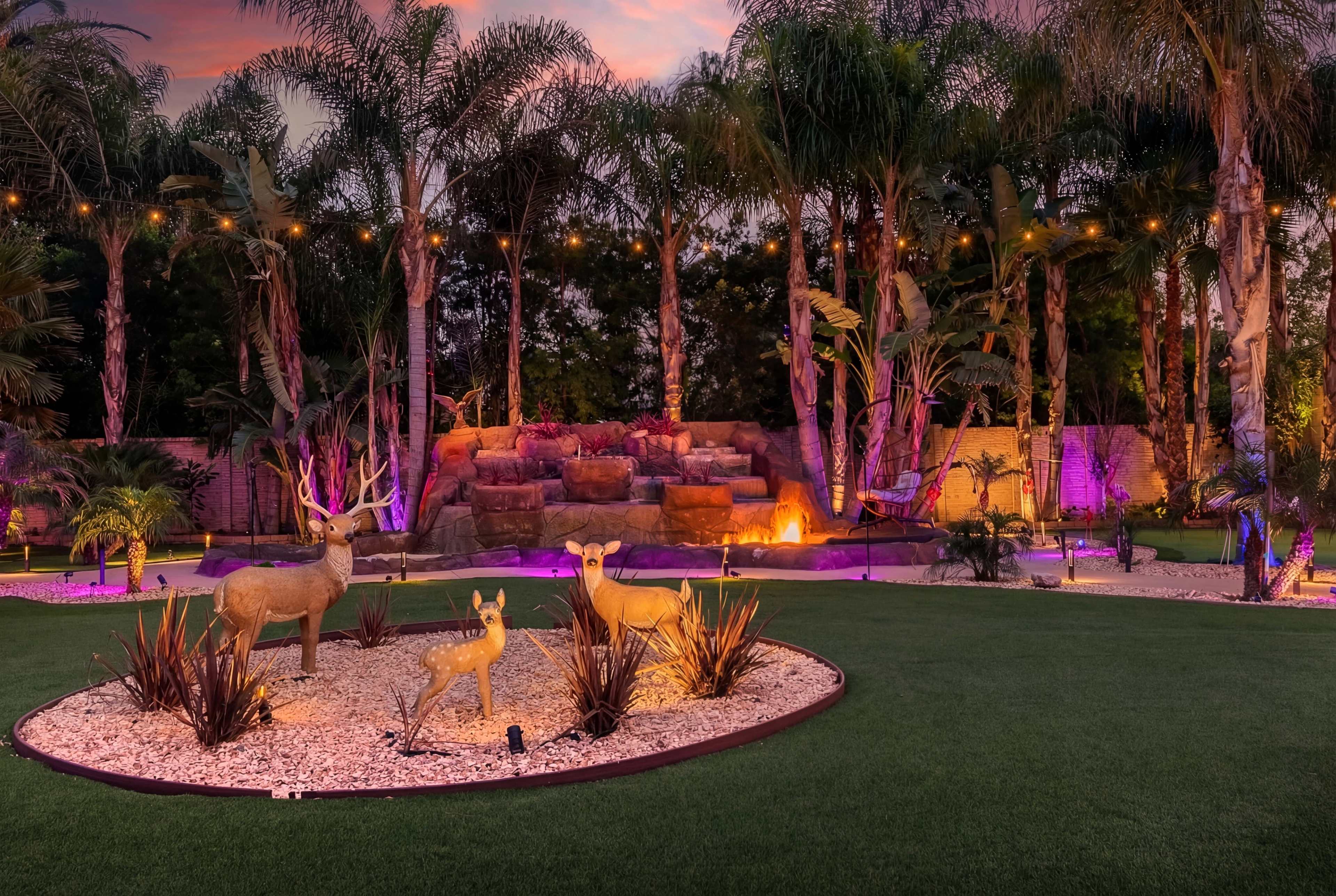The image shows a landscaped backyard featuring deer statues, surrounded by lush palm trees, decorative rocks, and vibrant lighting.