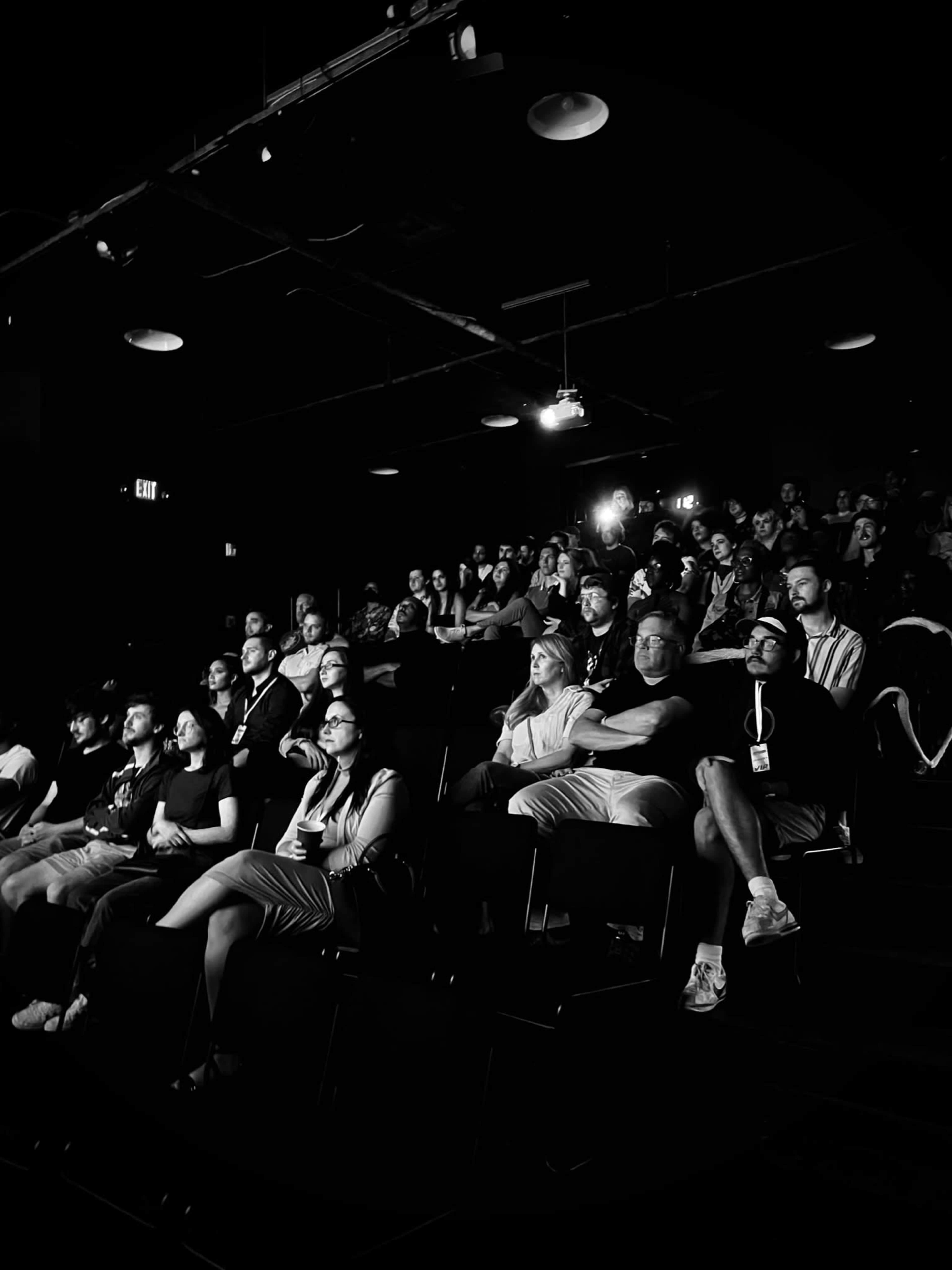 A group of people sits in a darkened theater, attentively watching a presentation on stage.