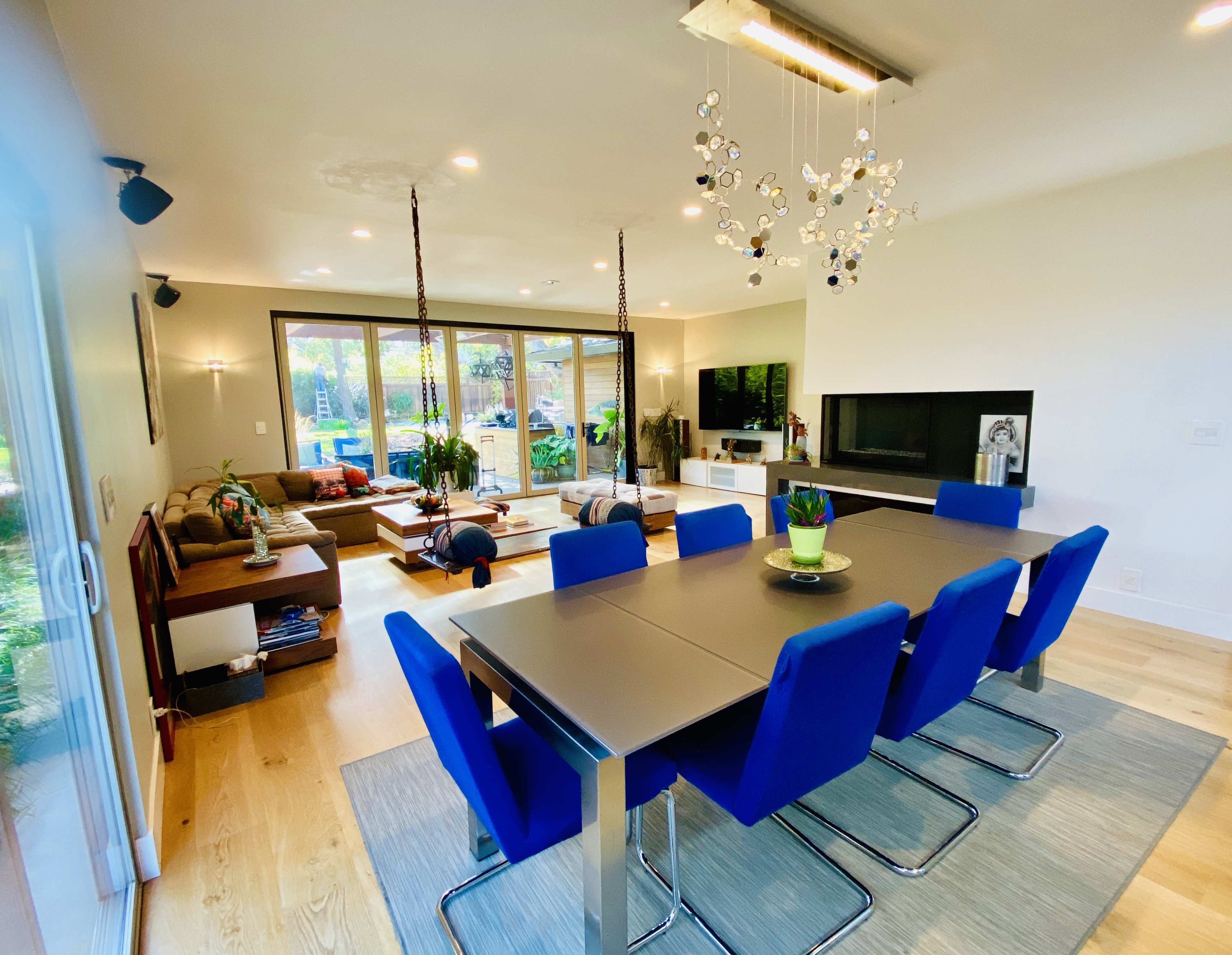 A modern dining area features a sleek table with blue chairs, a chandelier, and large windows overlooking a backyard with greenery.