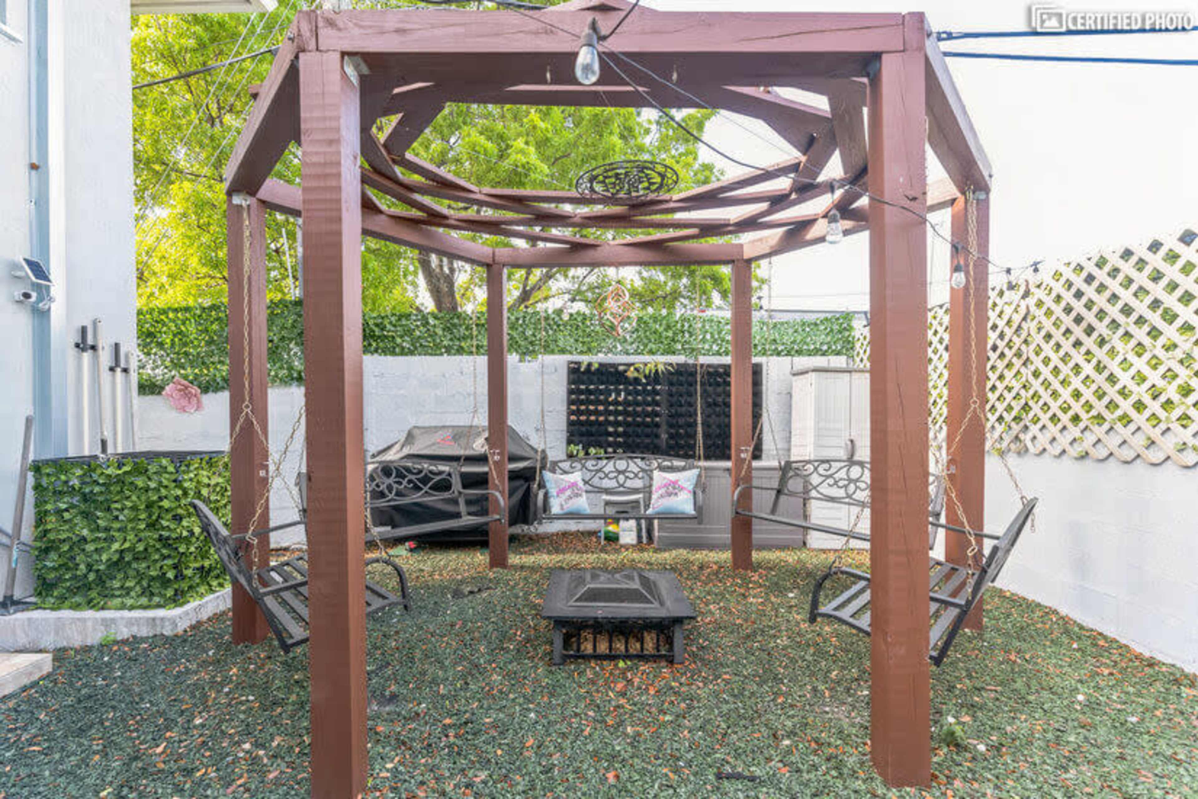 A wooden gazebo with swing seating is surrounded by greenery and outdoor furnishings in a spacious outdoor area.