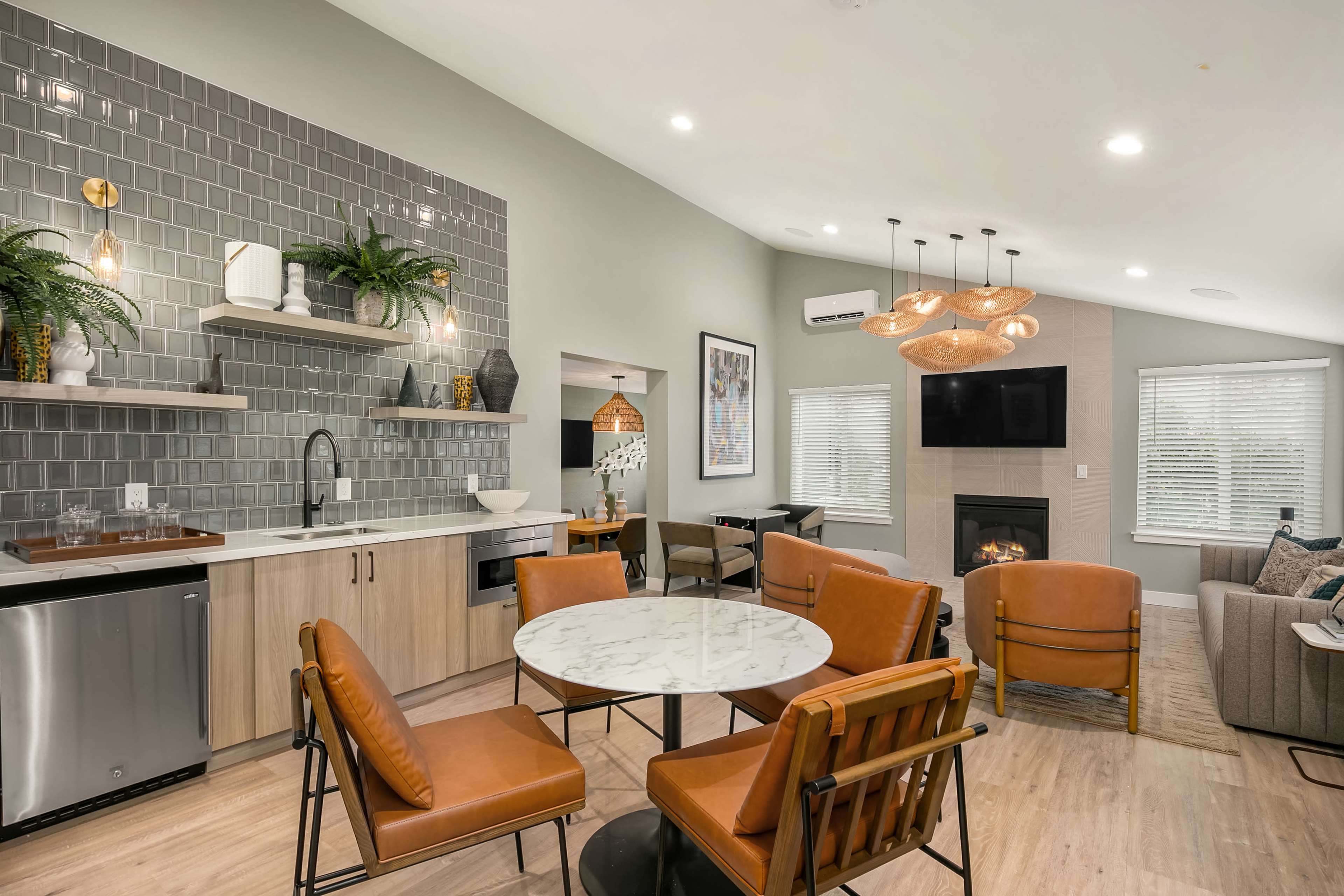 The image shows a modern living space featuring a dining area with a round marble table and brown chairs, a kitchenette with appliances, and a cozy seating area with a fireplace and a television.