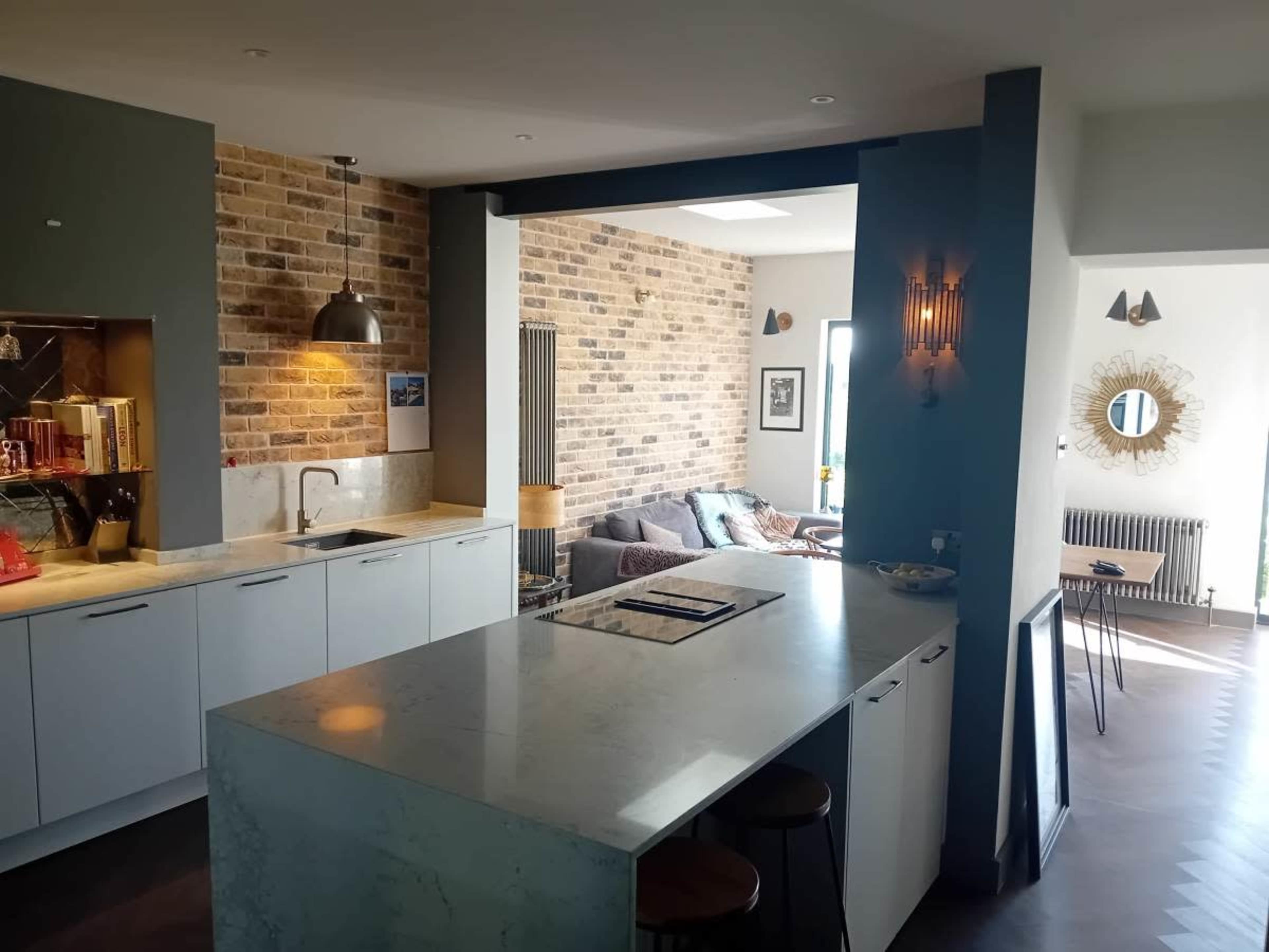 The image shows a modern kitchen featuring a marble island, white cabinetry, and a brick accent wall that connects to a living area with a couch and decorative mirror.
