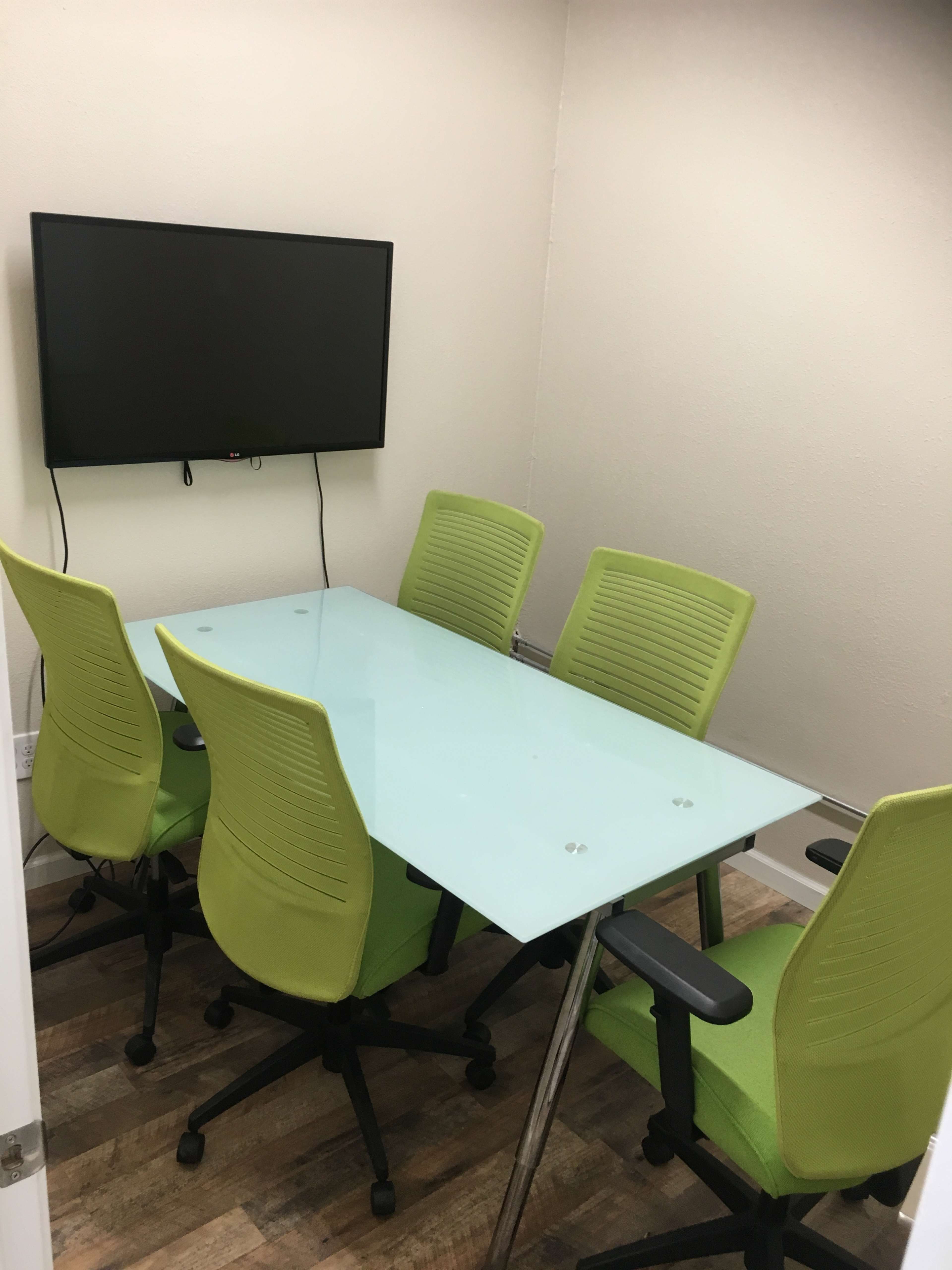 A small conference room features a glass table surrounded by five green office chairs and a wall-mounted television.