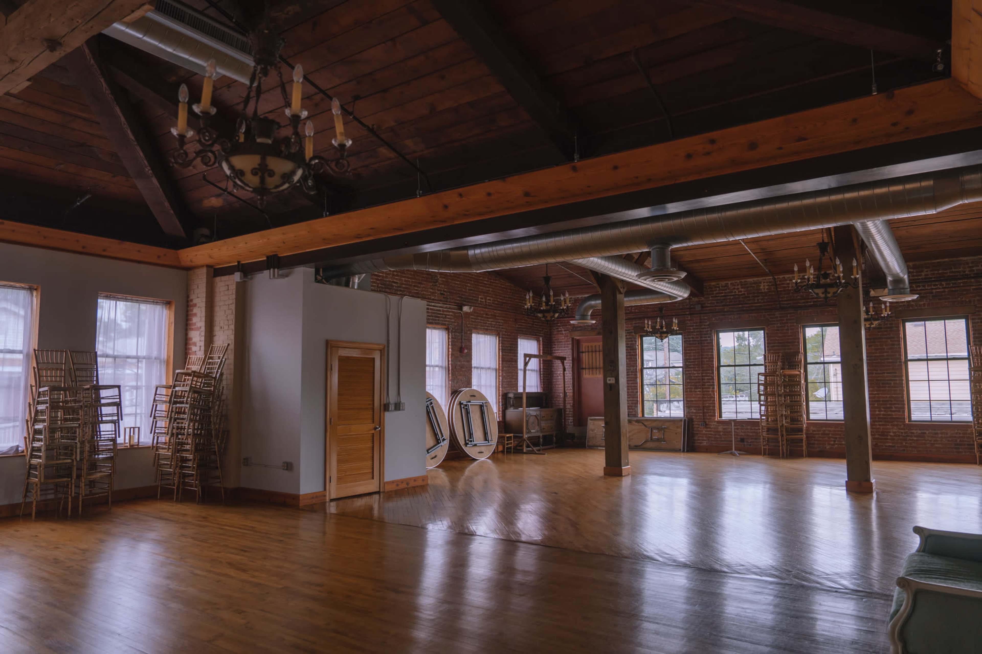 The image shows a large, open room with wooden floors, exposed brick walls, and tall windows, furnished with stacked chairs and a few decorative fixtures.