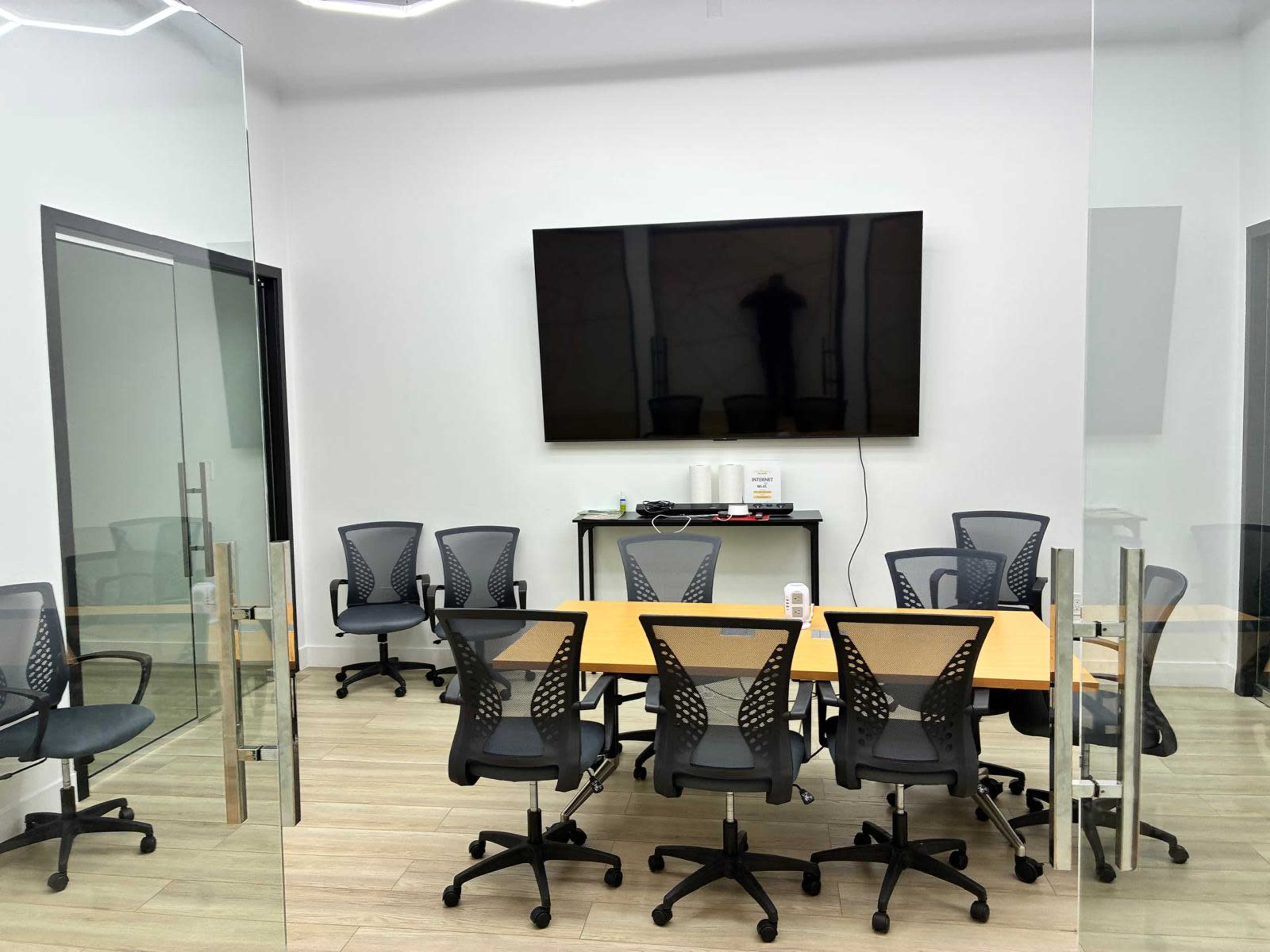 The image shows a modern conference room with a large wall-mounted TV, a rectangular table surrounded by ergonomic chairs, and glass walls.