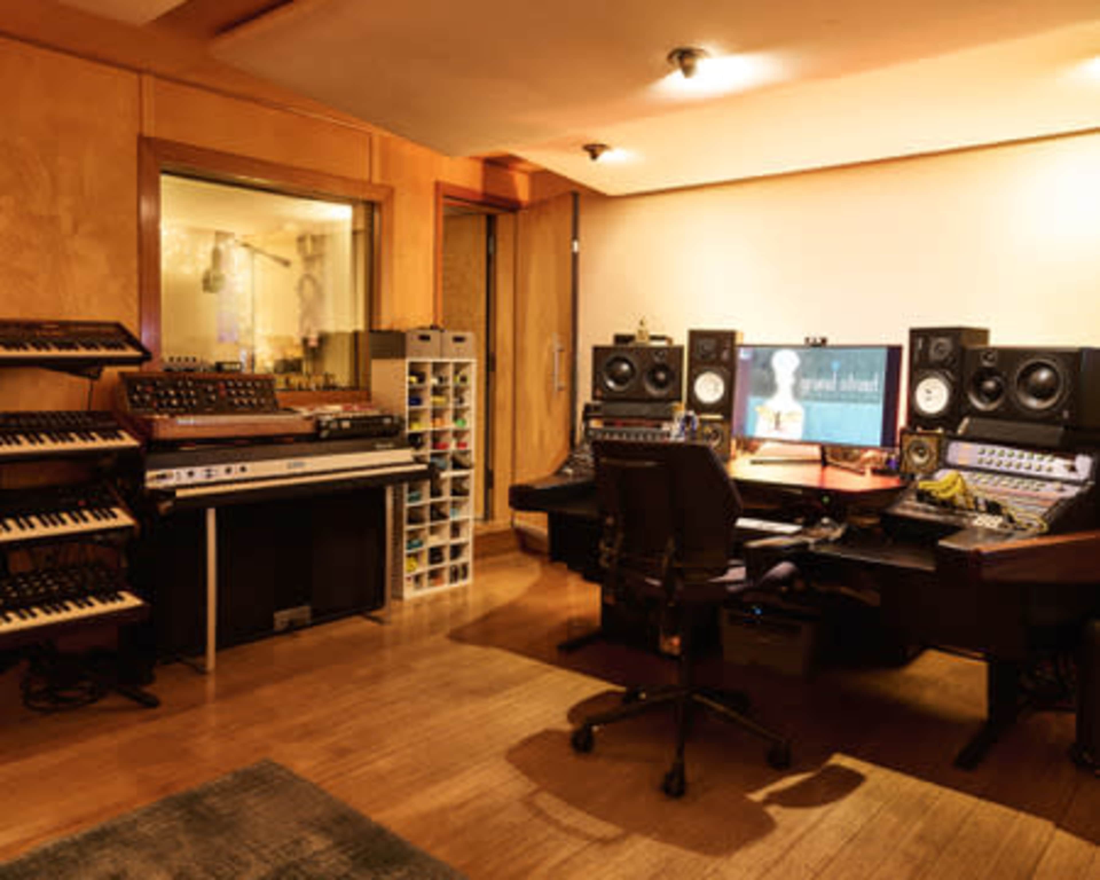 The image shows a music studio with various synthesizers, speakers, and a large computer monitor displaying audio software.