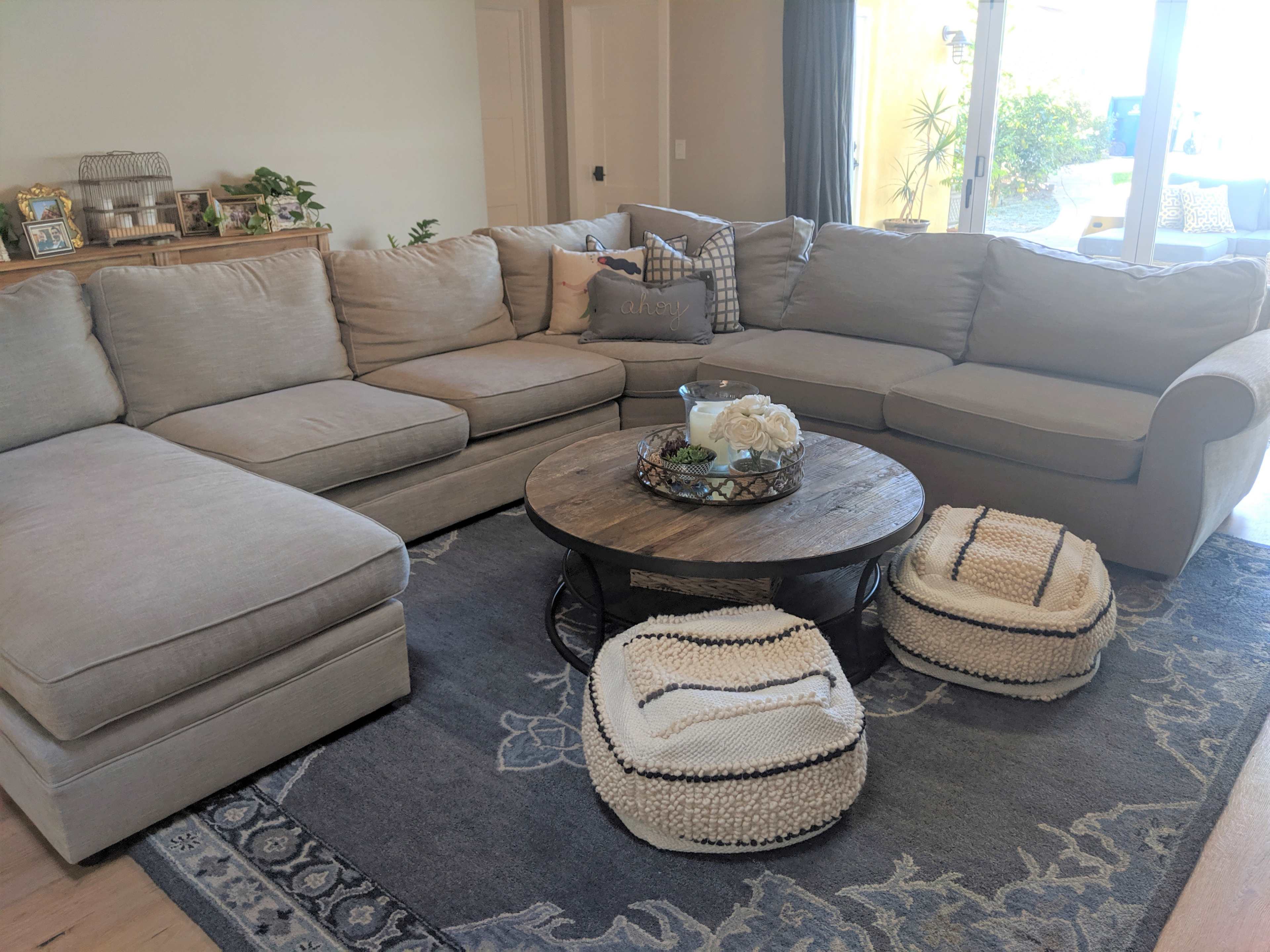 A sectional couch is positioned around a round wooden coffee table, with decorative items and plants in a well-lit living room.