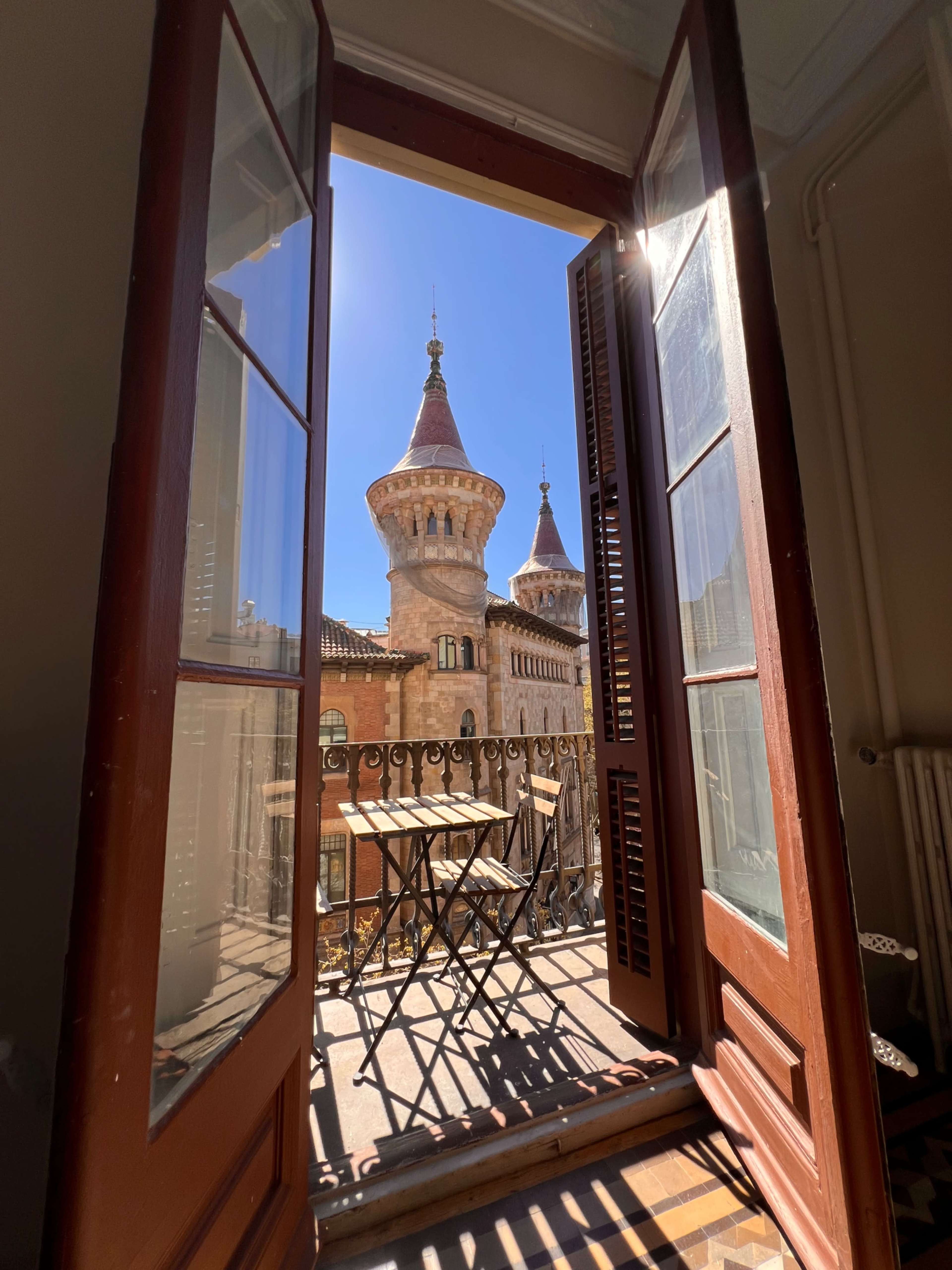 The image shows an open balcony door with a view of a tower featuring spires on a clear day.