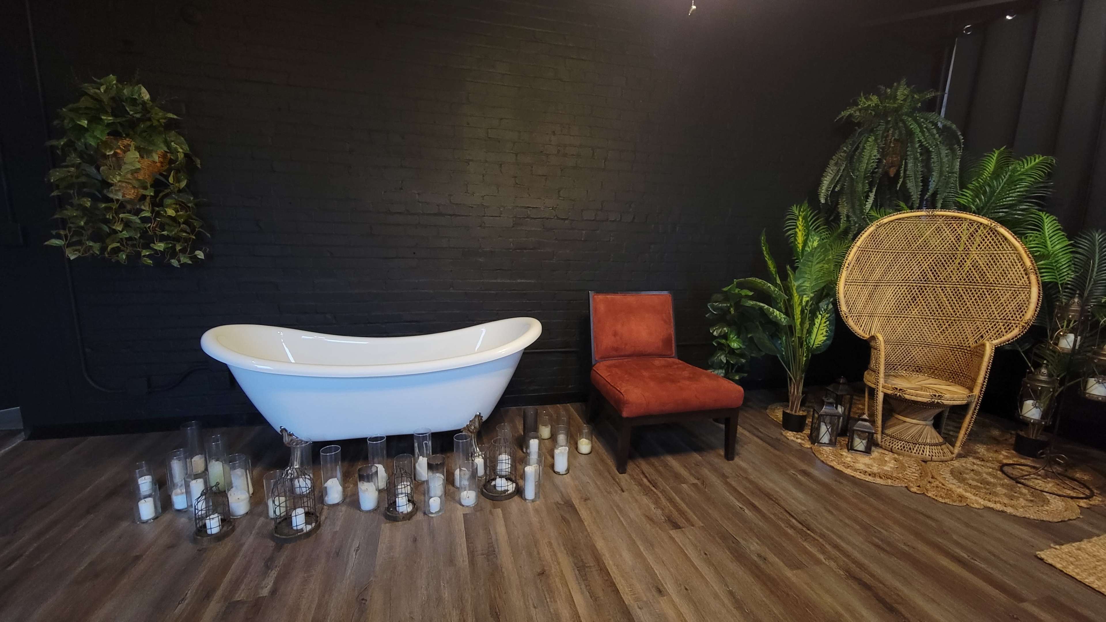 A minimalist bathroom interior with a freestanding white bathtub, a red chair, and various candles arranged on the floor, accompanied by greenery and a rattan chair.