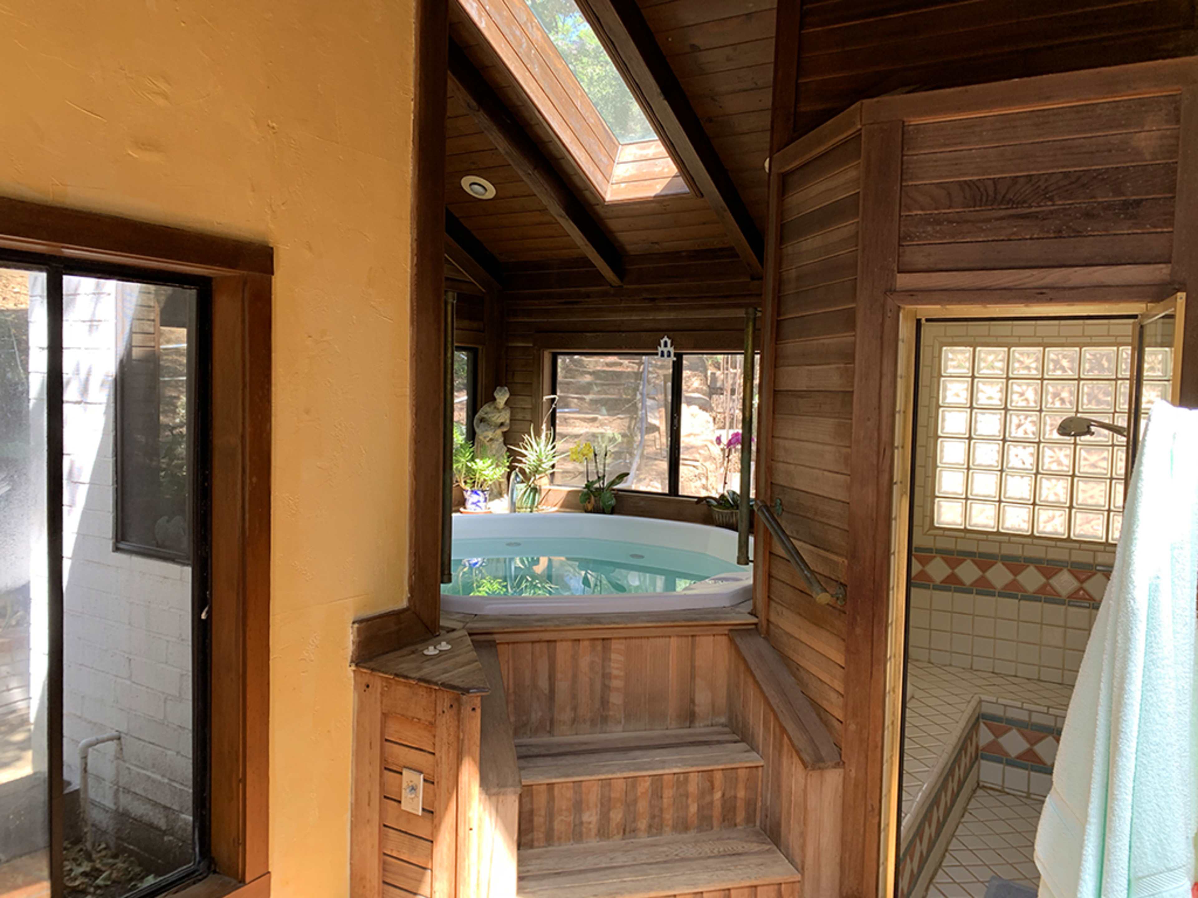 The image shows a spa-like area with a circular tub, large windows, and wooden walls, connected to a tiled shower space.
