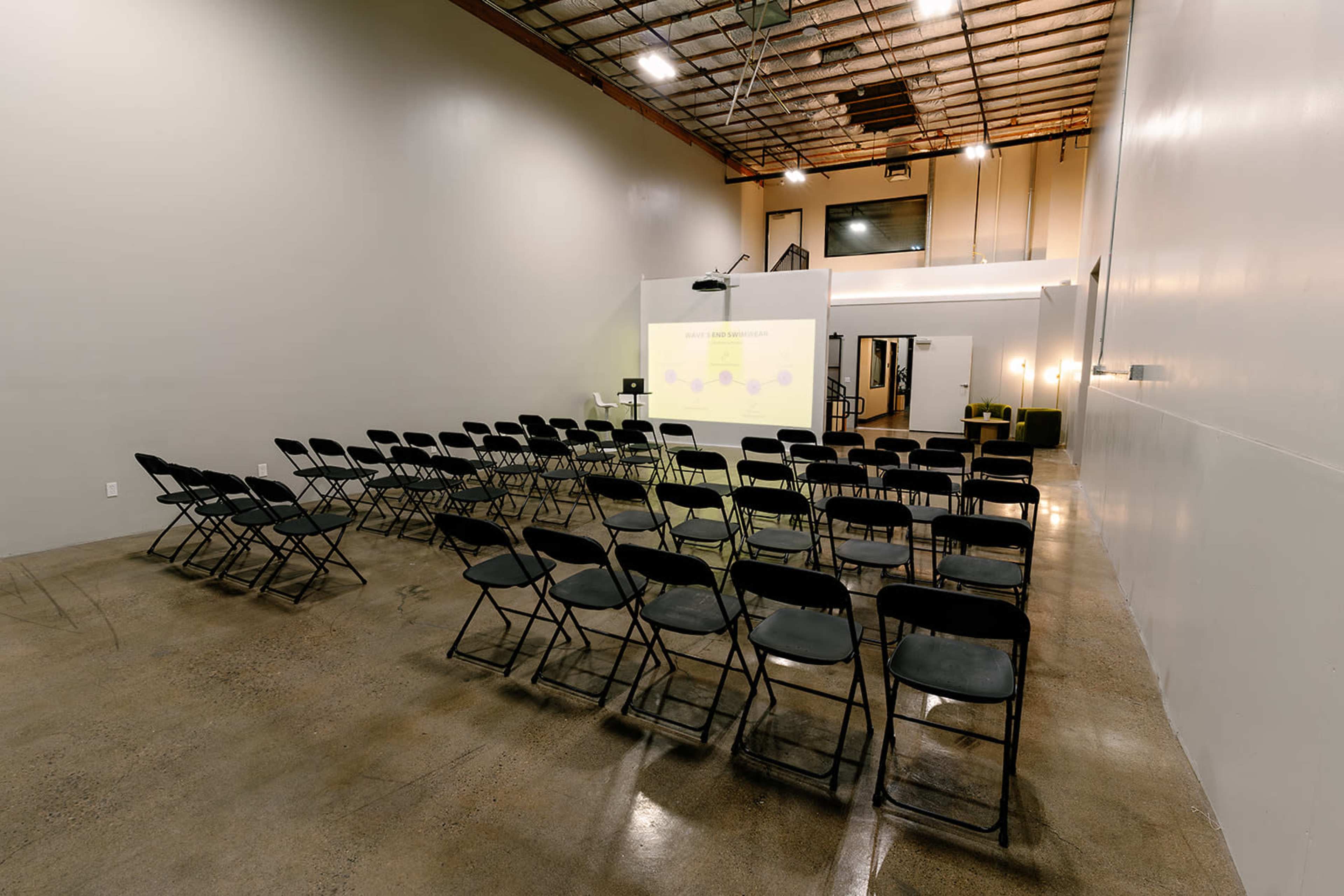 A spacious room features rows of black folding chairs facing a projector screen at the front.