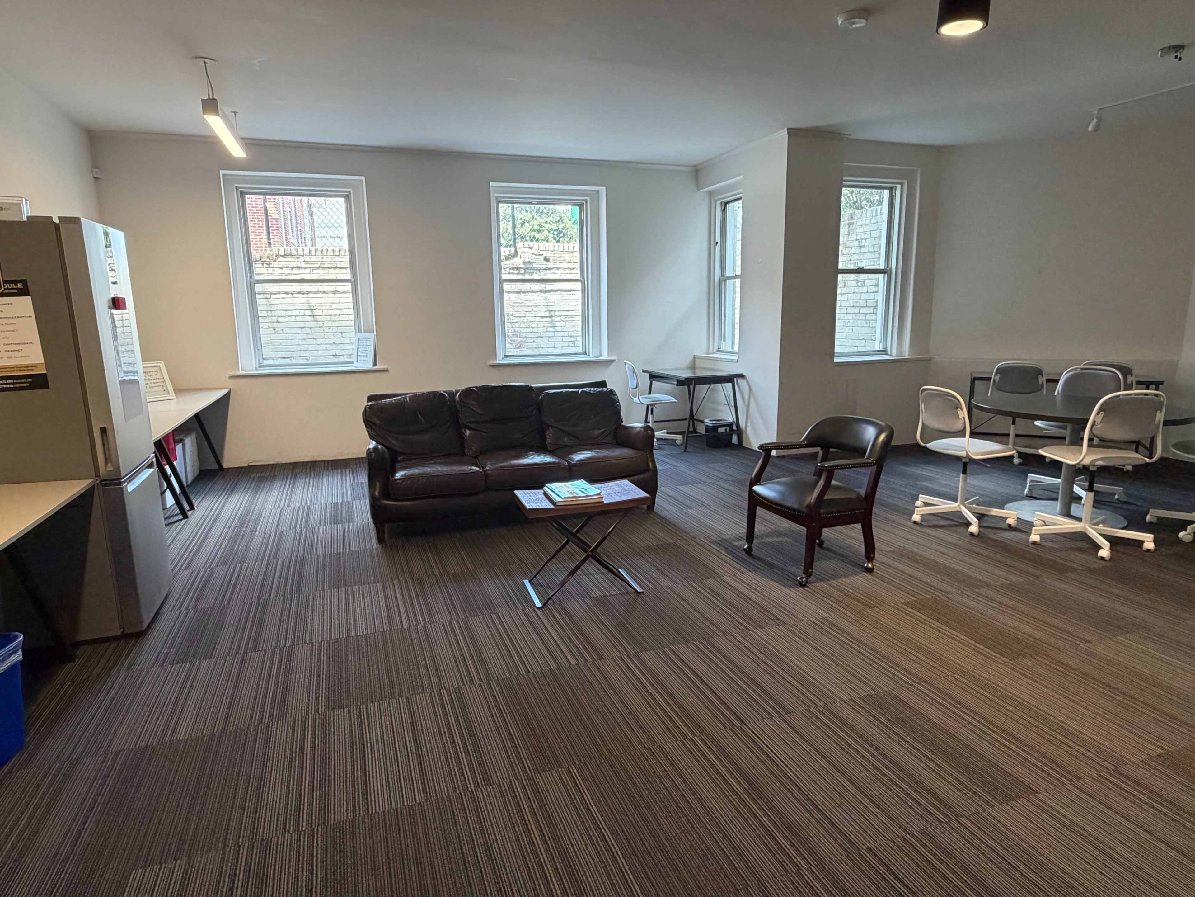 The image shows a spacious room with a brown couch, a coffee table, a chair, a desk with chairs, and large windows providing natural light.