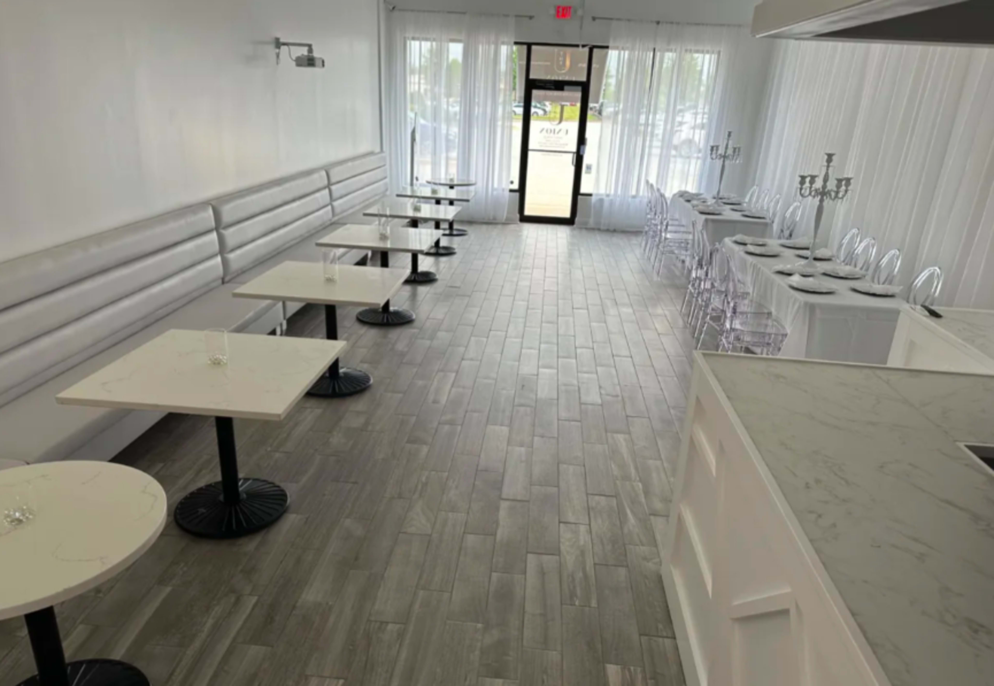The image shows a spacious, empty dining area with white walls, several marble-top tables with black bases, and large windows leading to an entrance.