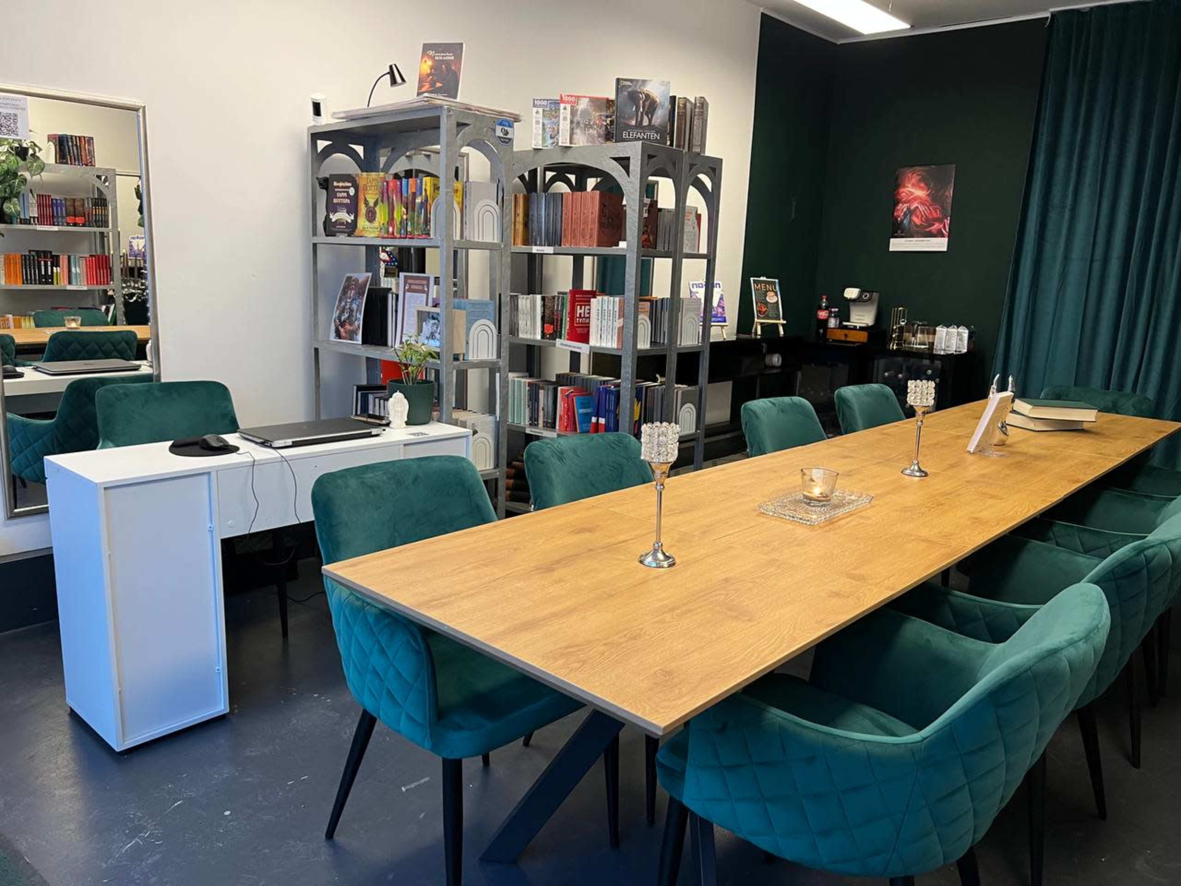 The image shows a modern meeting room with a long wooden table surrounded by green chairs, shelves filled with books, and a mirror on one wall.