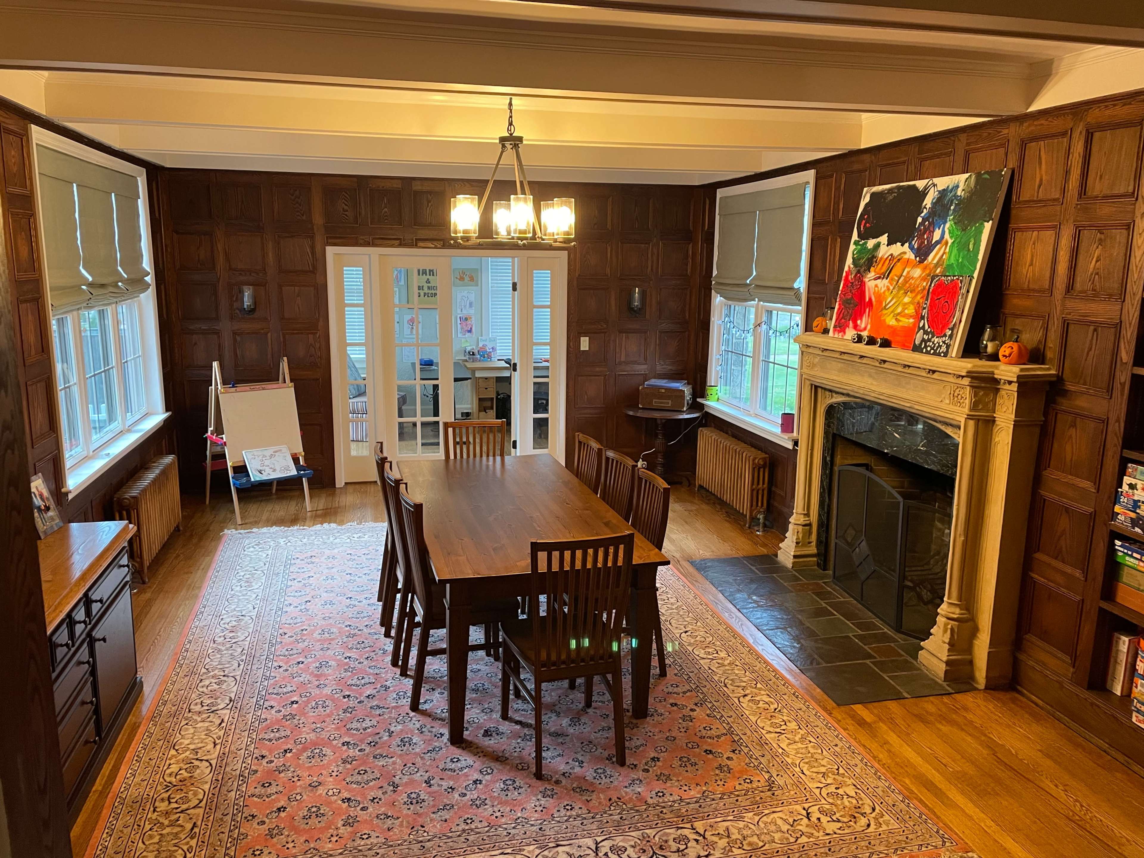 The image shows a spacious dining room with a wooden table and chairs, a fireplace, and large windows, illuminated by a chandelier.