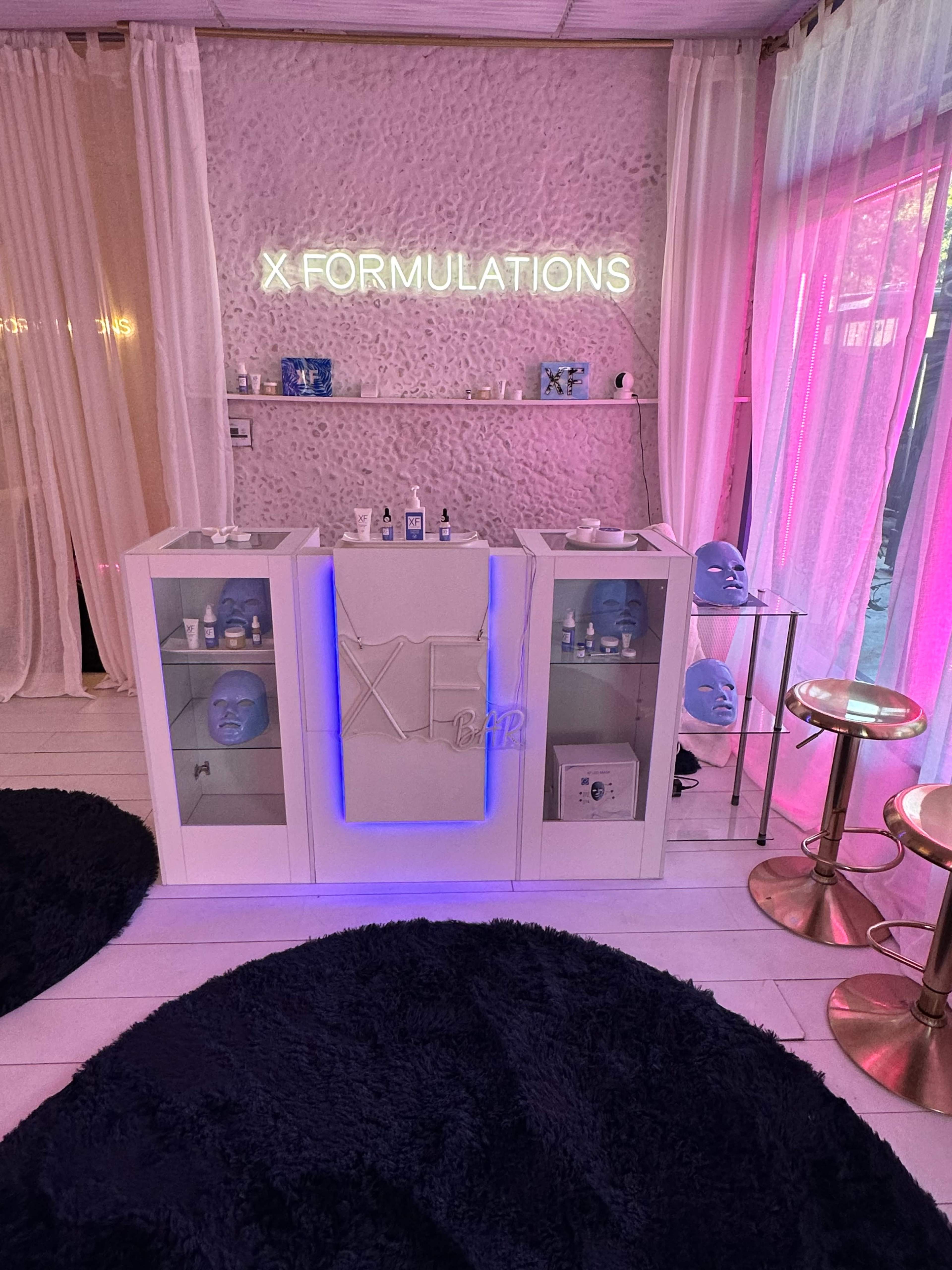 The image shows a modern skincare treatment area featuring a sleek white counter with various skincare products and blue-faced treatment masks, illuminated by a neon sign that reads "X FORMULATIONS."