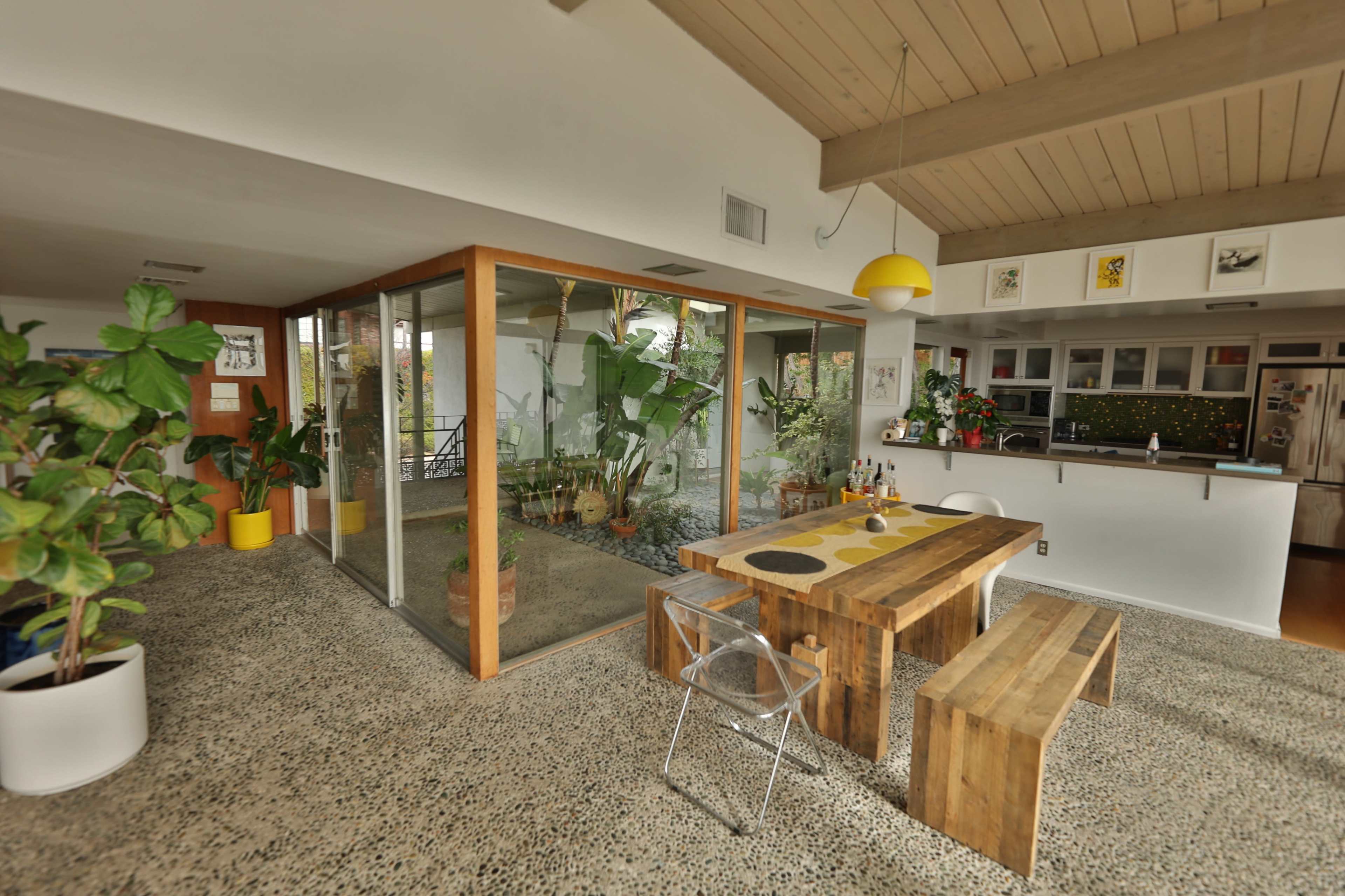 The image shows a modern open-concept kitchen and dining area with a wooden table and benches, surrounded by glass walls and indoor plants.