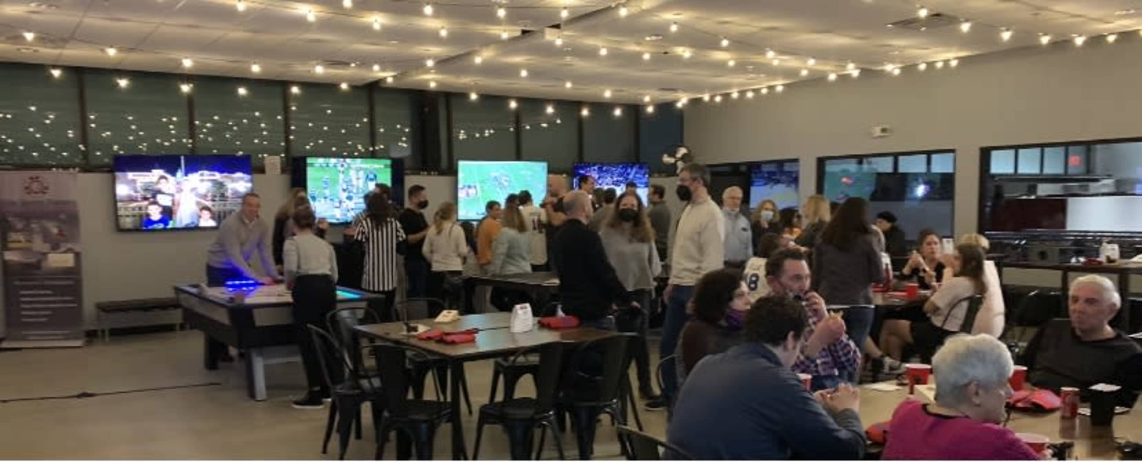 The scene shows a crowded indoor entertainment venue with large screens displaying games and groups of people socializing at tables.