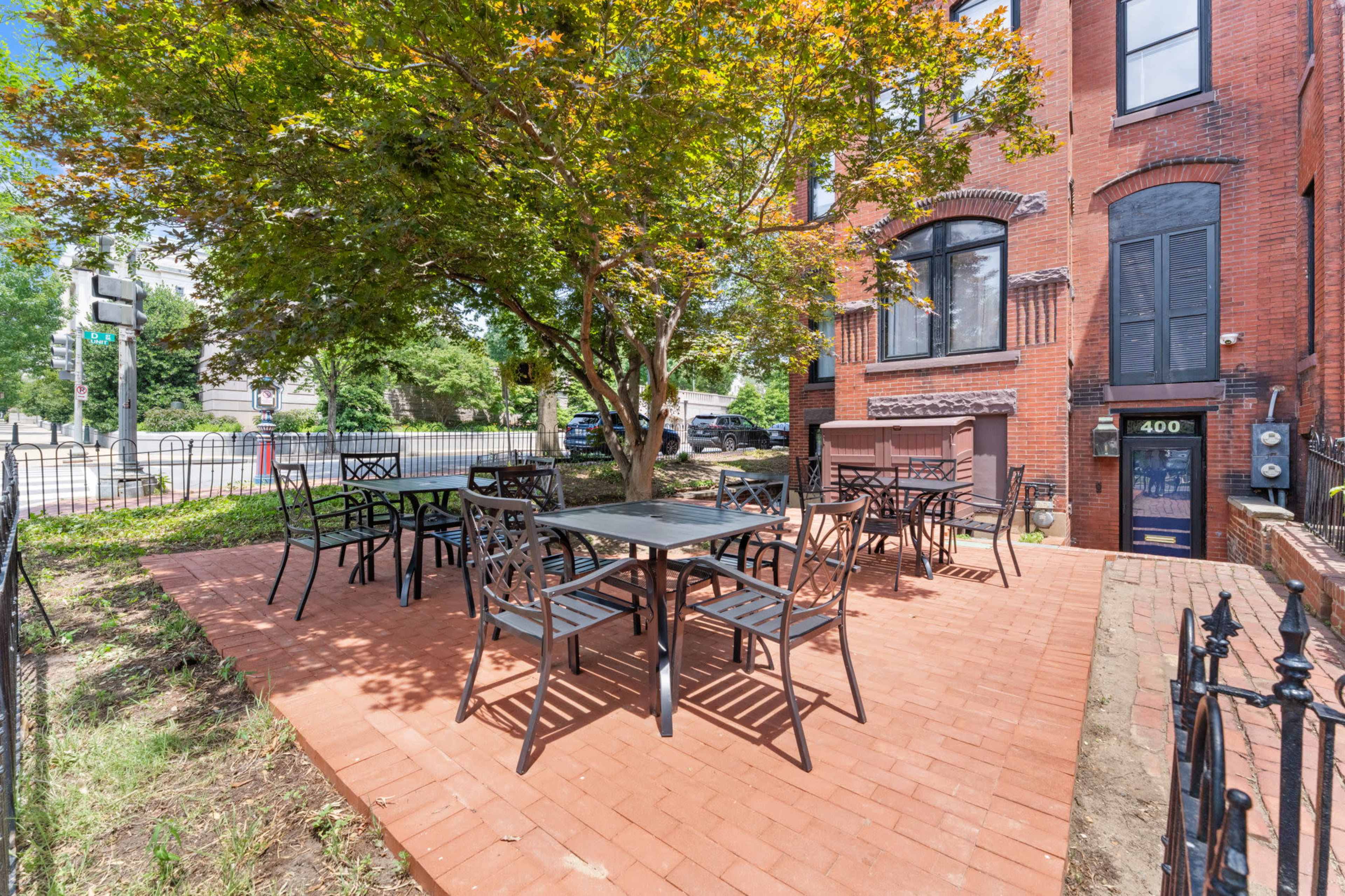 The image shows a brick patio area with black metal tables and chairs arranged around a central tree, adjacent to a red brick building.