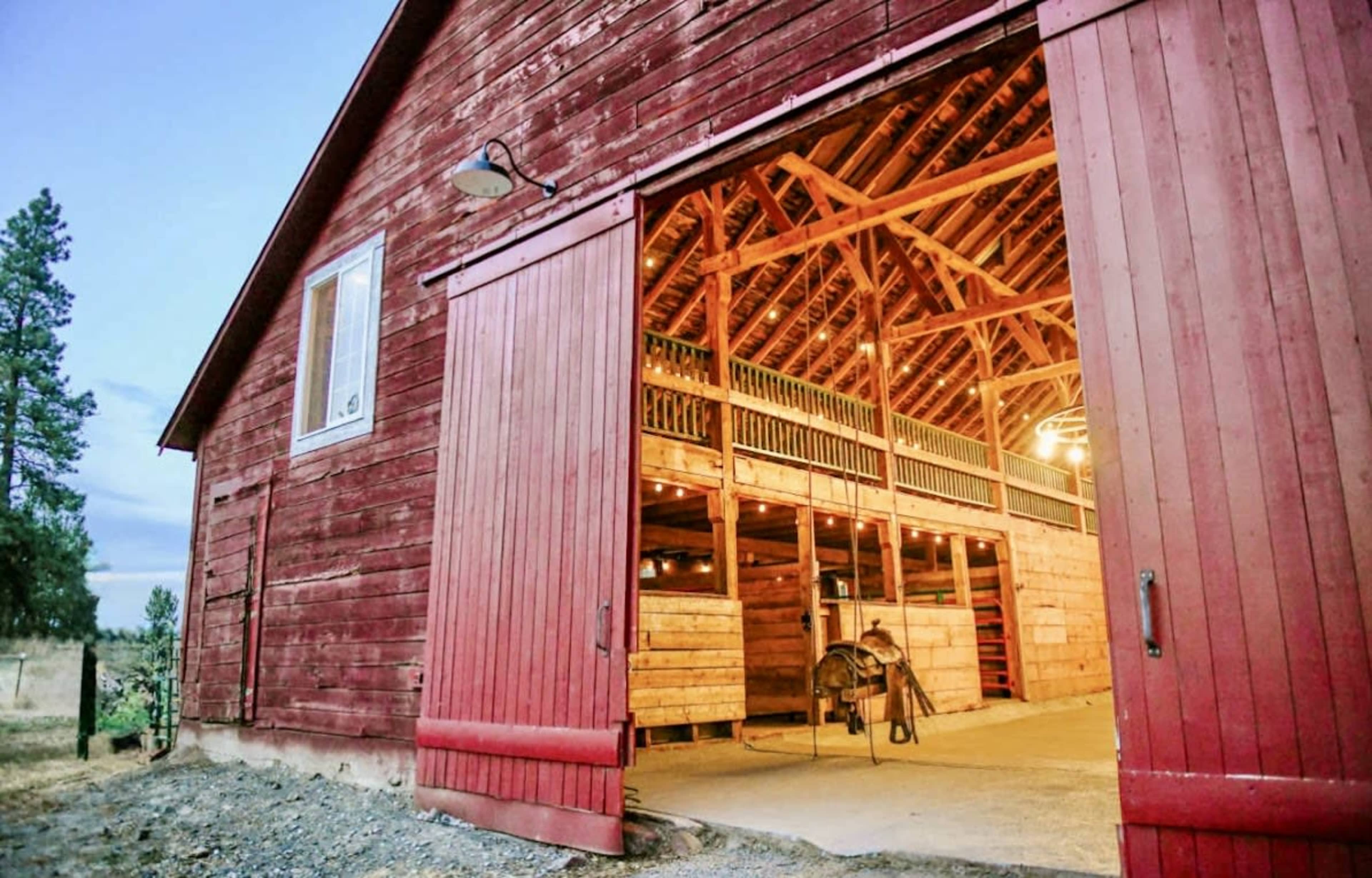 The image shows a rustic red barn with open double doors revealing a well-lit interior featuring wooden stalls and beams.