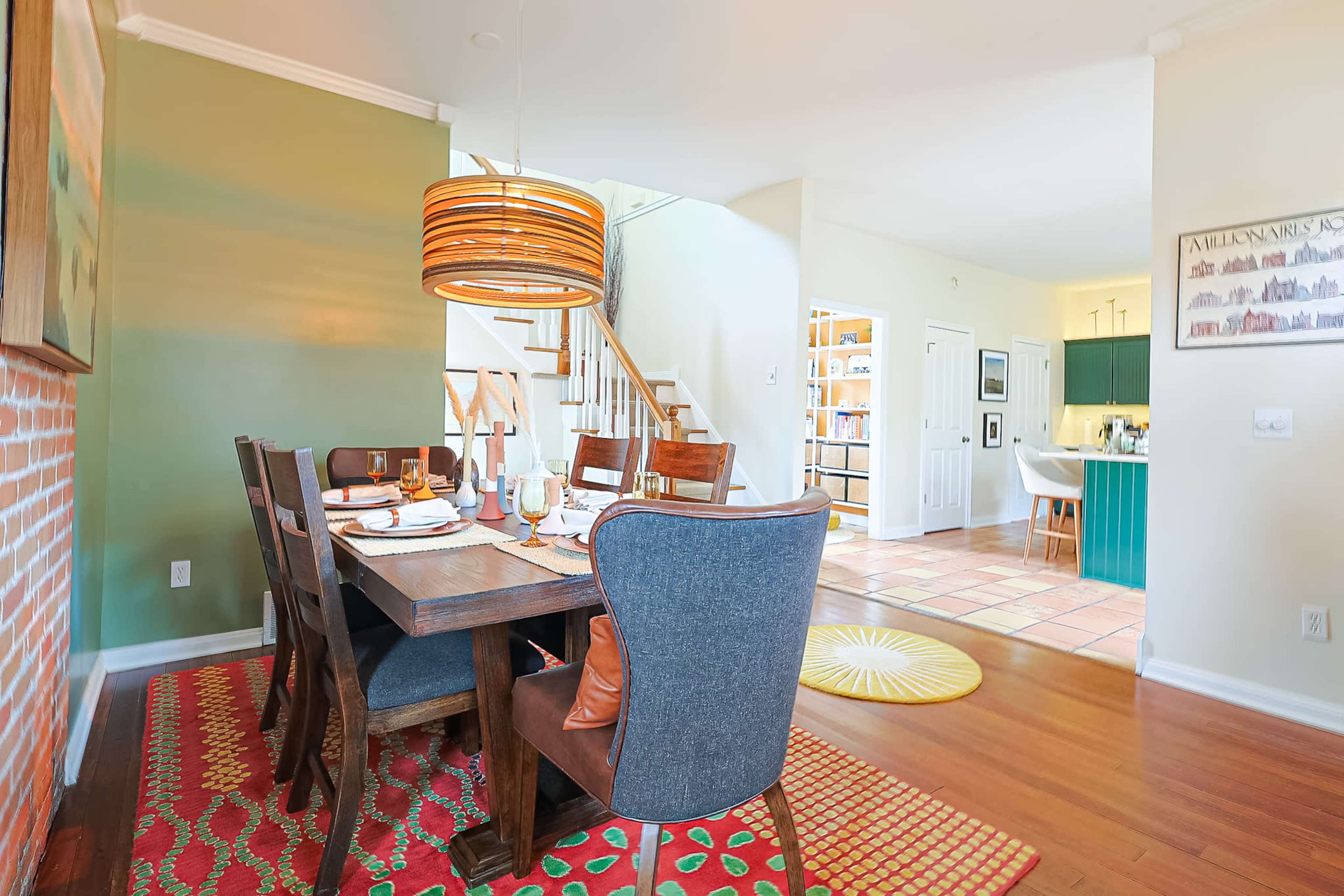 The image shows a dining room with a wooden table set for a meal, surrounded by upholstered chairs, and a staircase leading to another level in the background.