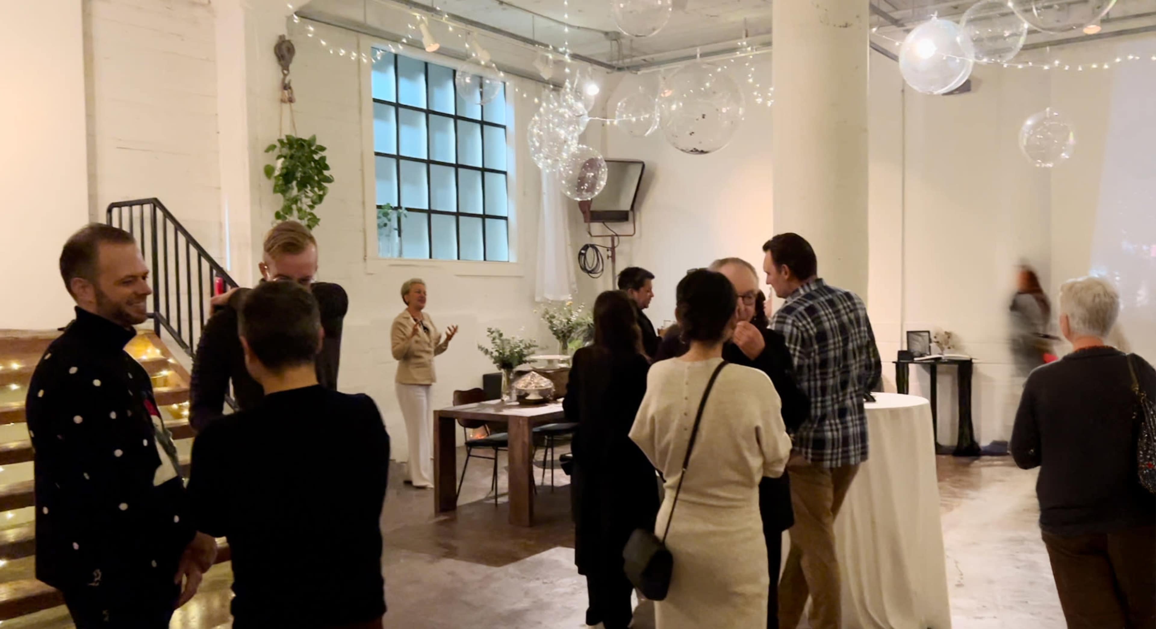 A group of people converse and mingle in a well-lit event space adorned with string lights and hanging decorations.