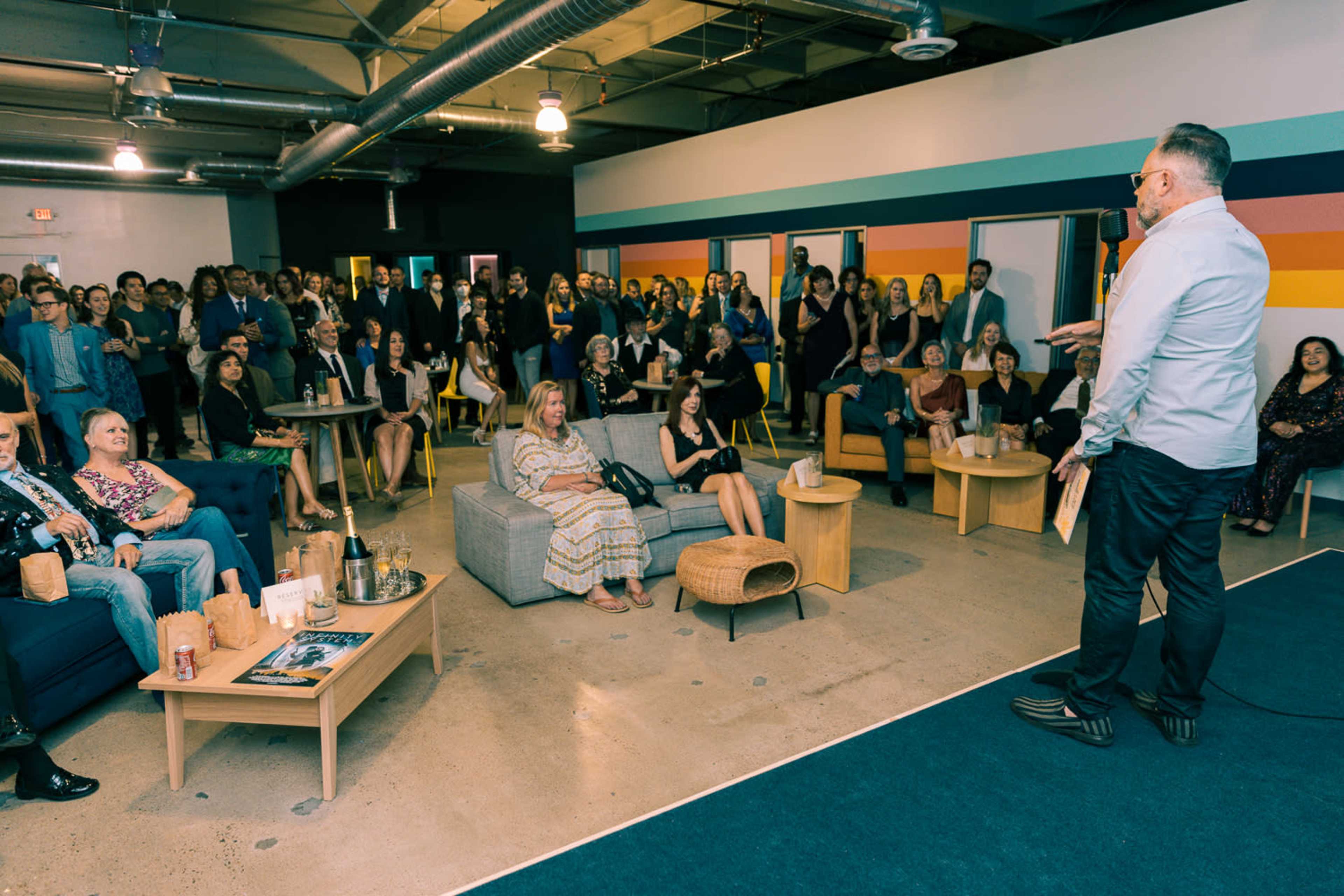 A speaker addresses an audience gathered in a modern lounge space with seating areas and colorful wall stripes.