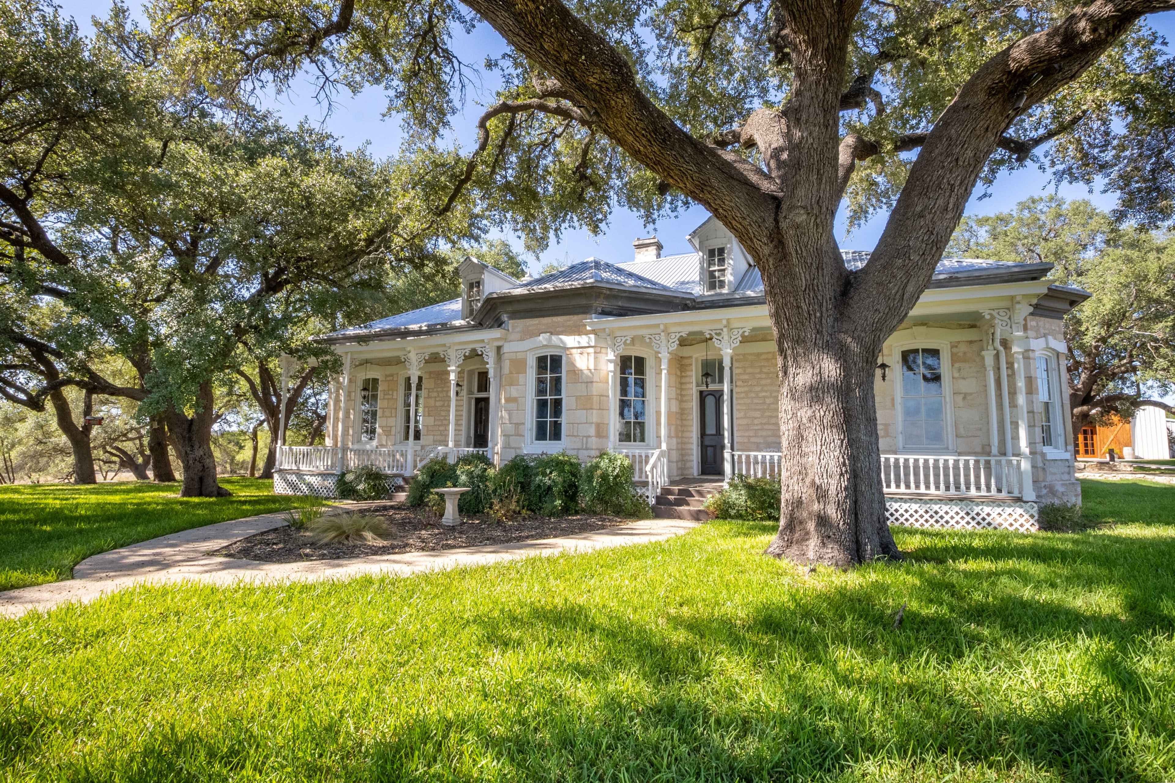 A historic house with a wraparound porch is surrounded by large trees and green grass.