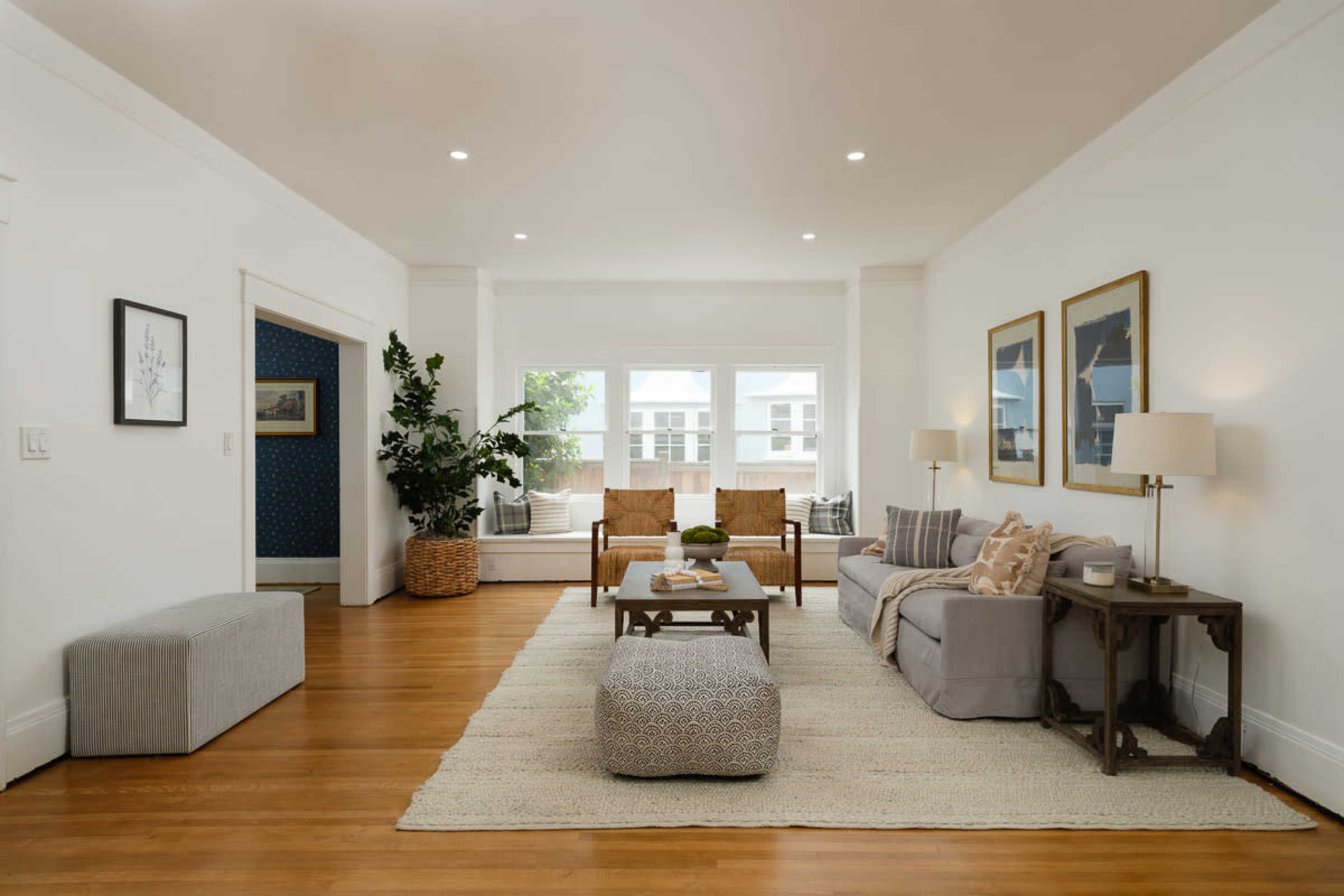The image shows a spacious living room featuring a gray sofa, two wooden armchairs, a coffee table, and large windows allowing natural light to enter.