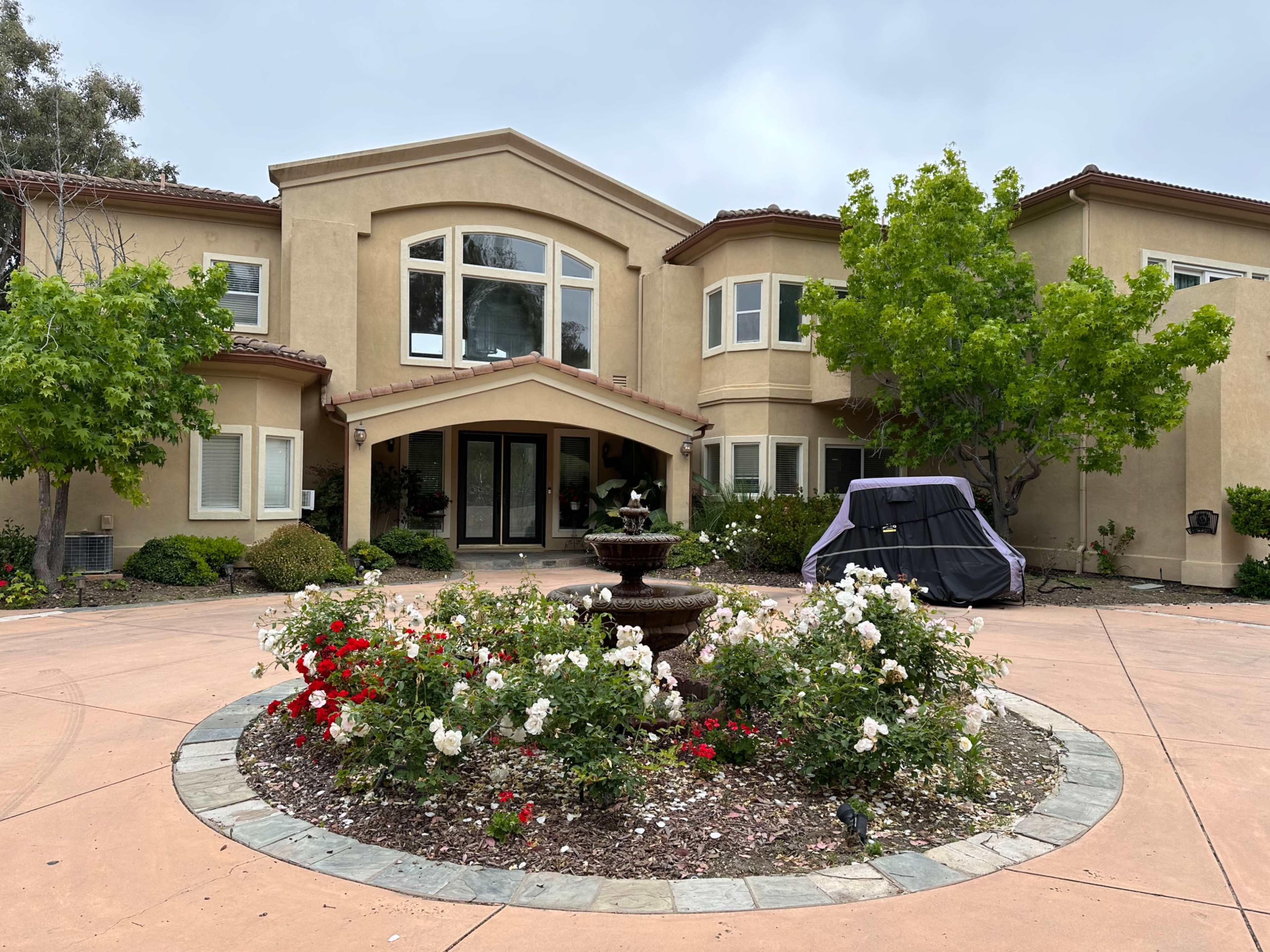 A large, two-story house with a circular garden and a fountain features blooming rose bushes in front.