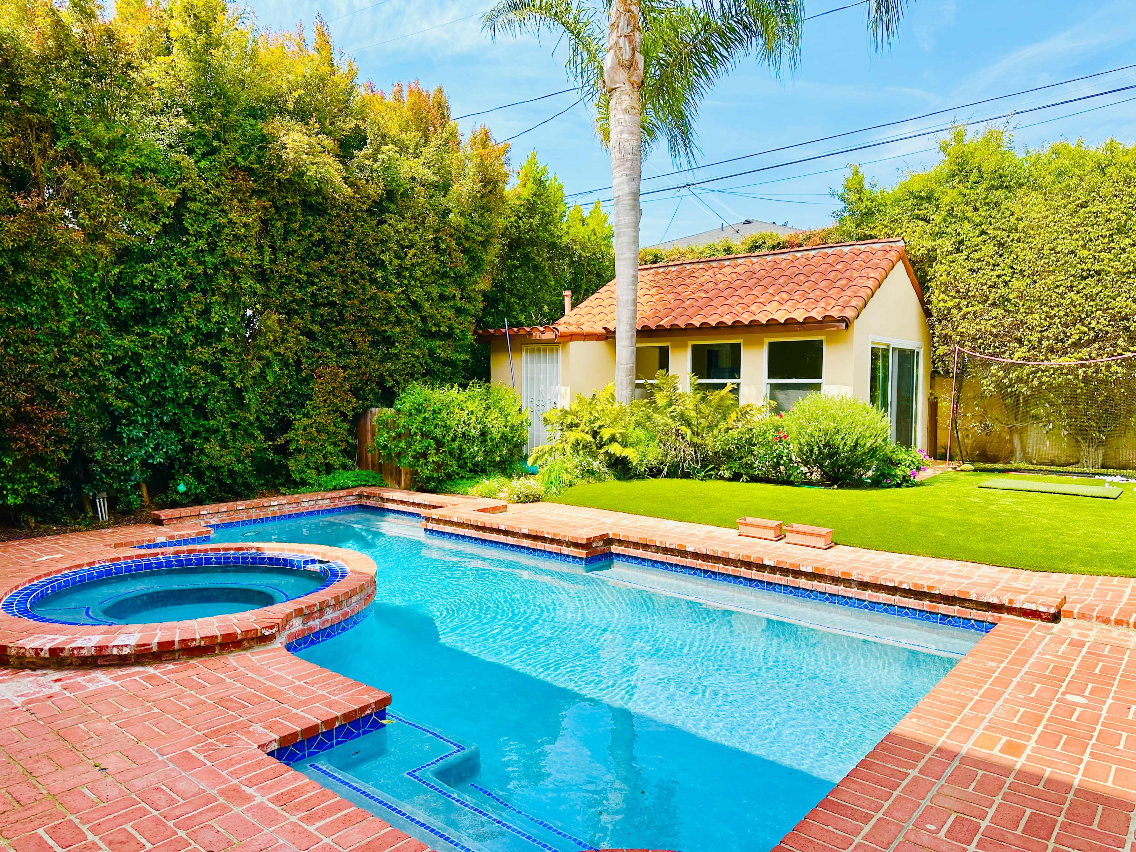 The scene features a backyard with a blue swimming pool, a spa area, and a small house surrounded by greenery.