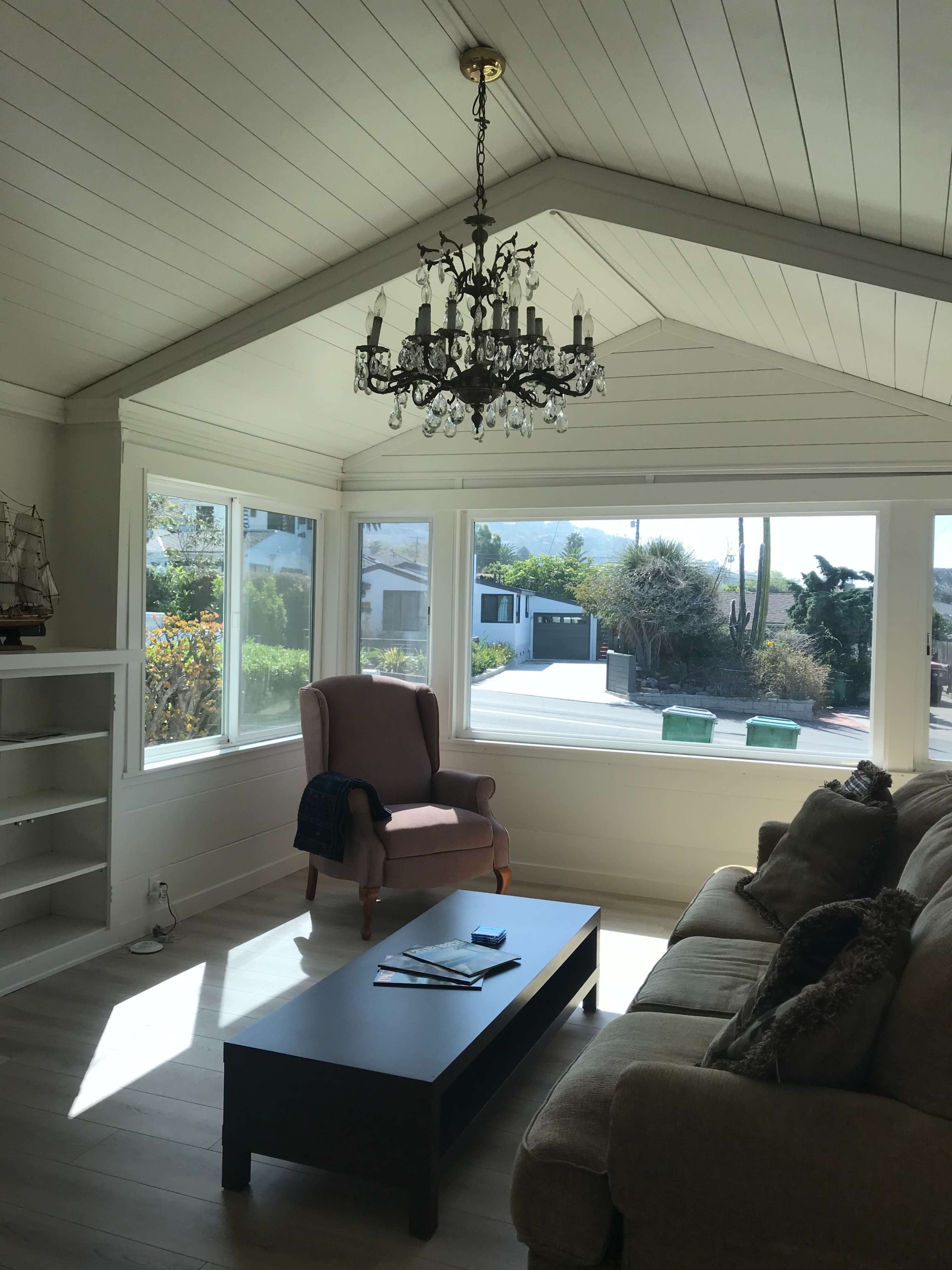 A sunlit living room with a chandelier, large windows, a pink armchair, a sofa, and a coffee table.