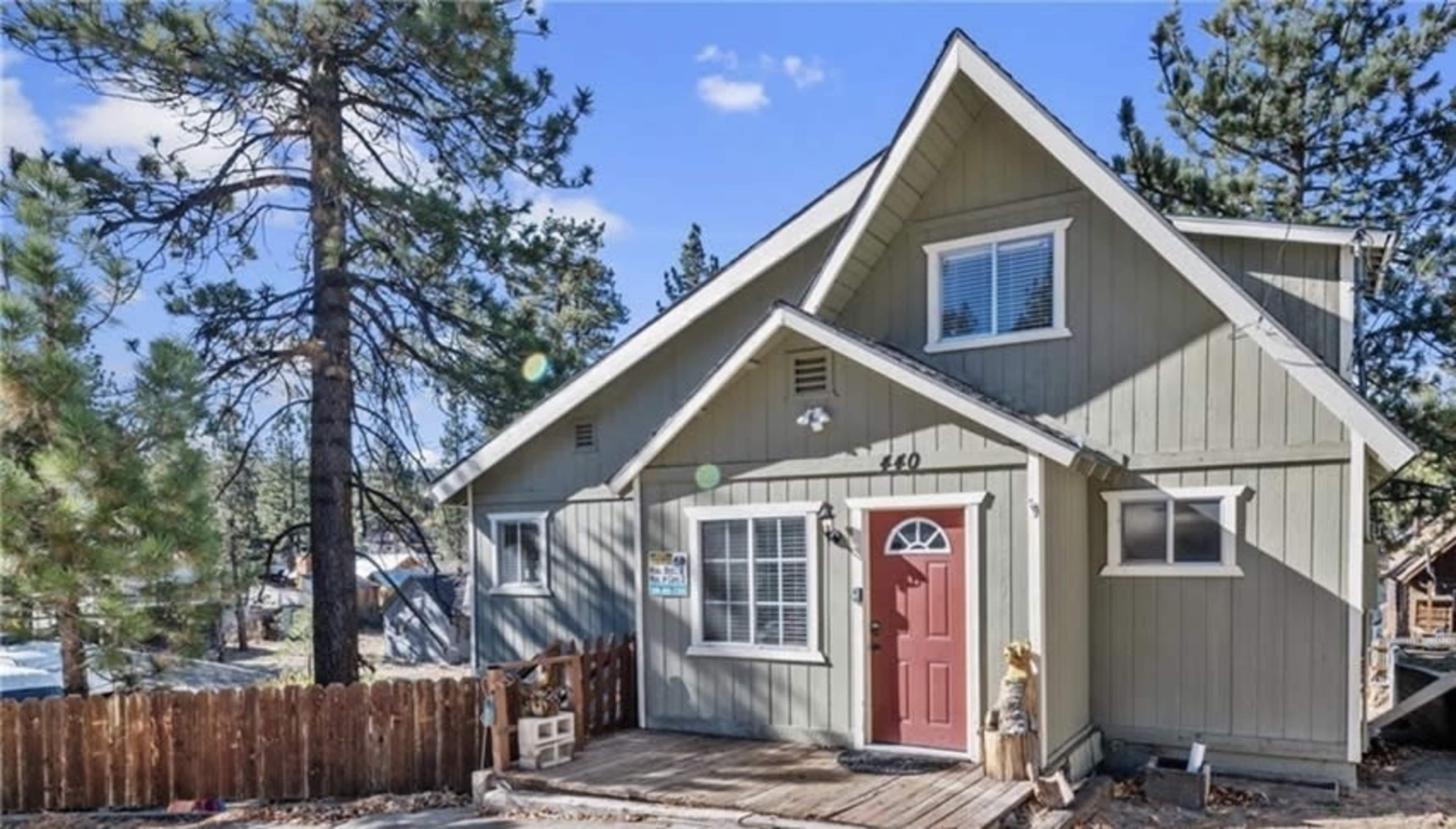 A green, two-story house with a red door and a wooden porch is surrounded by tall pine trees.
