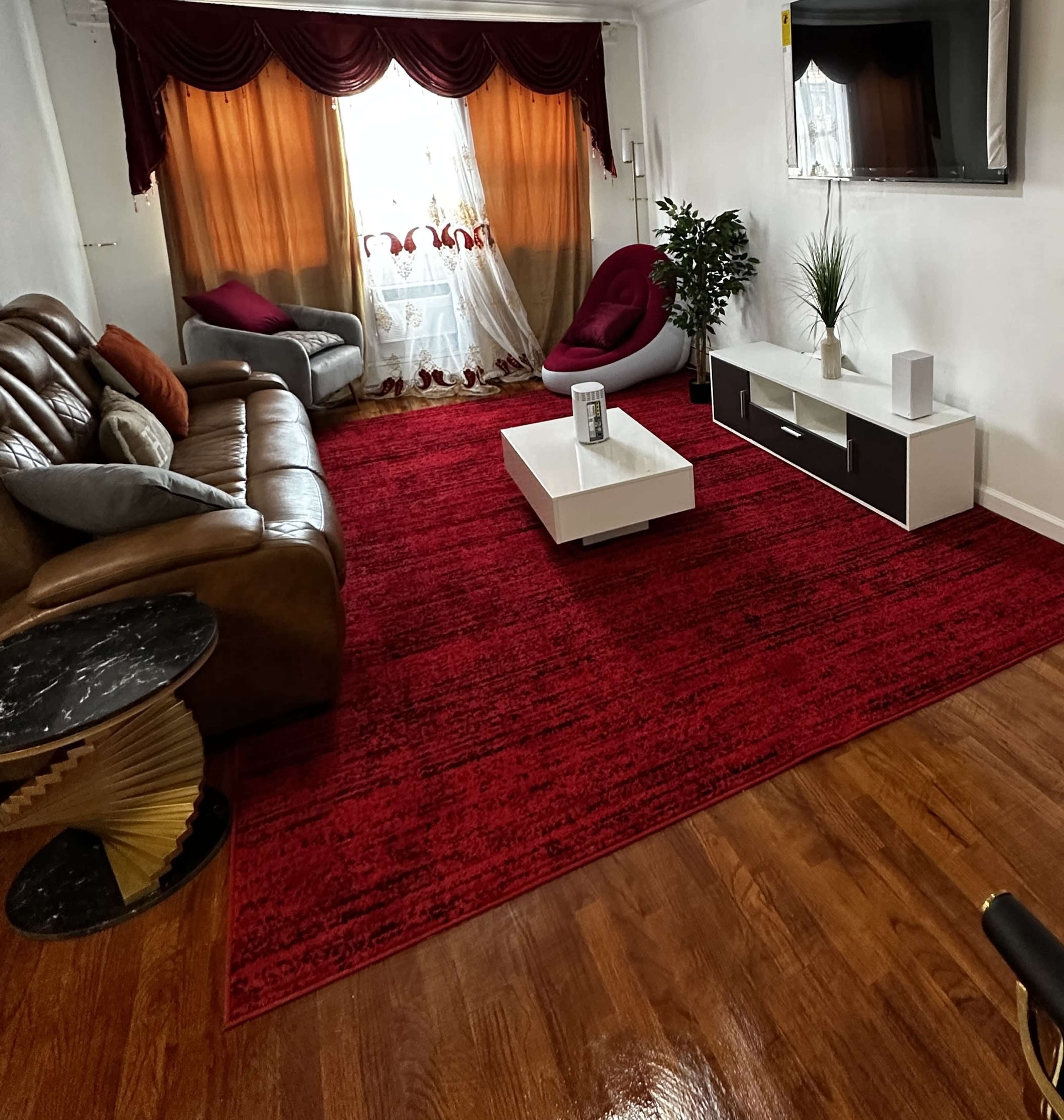 A living room with a red carpet, a brown leather sofa, a white coffee table, and large windows draped with orange curtains.