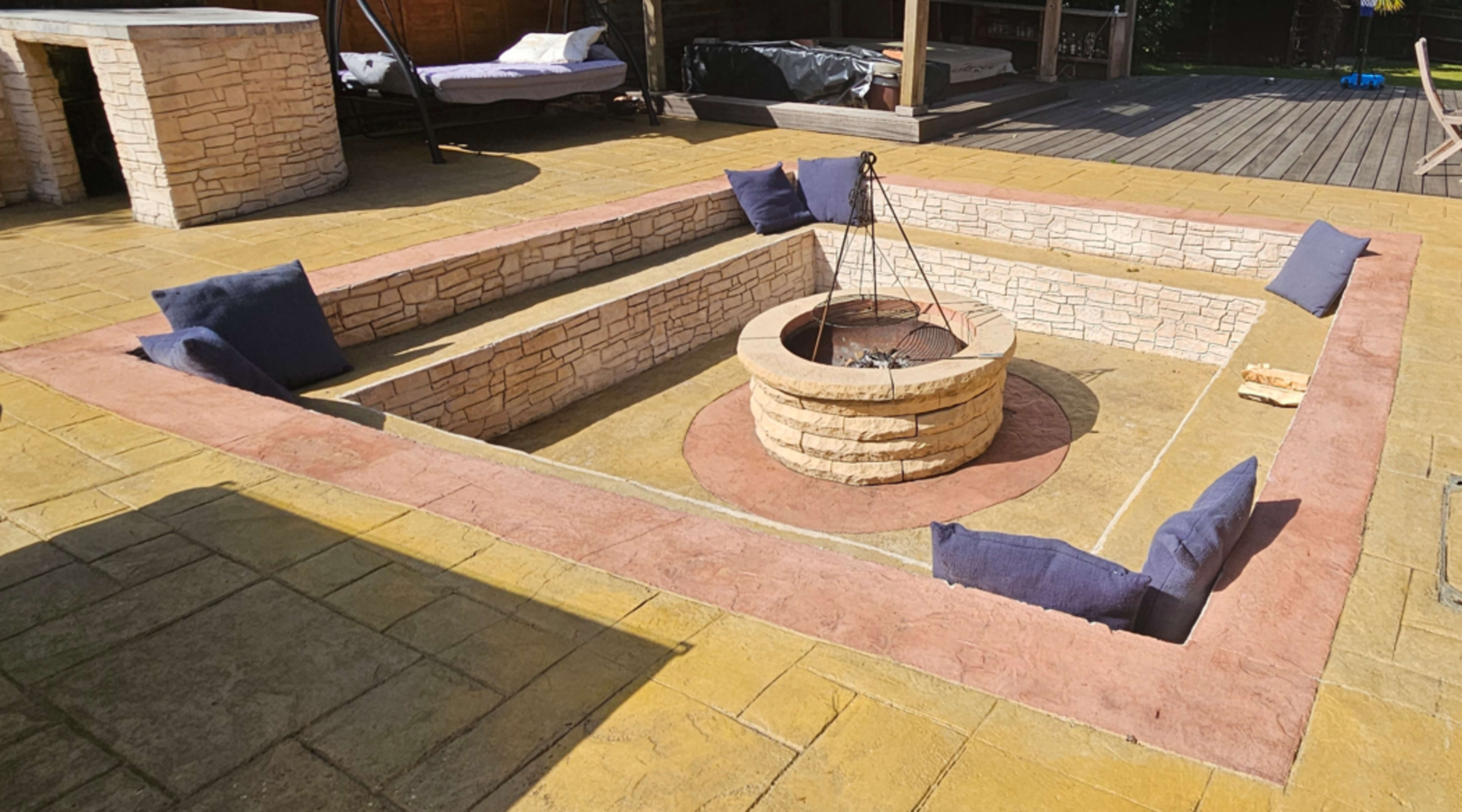 The image shows a stone fire pit surrounded by a rectangular seating area with blue cushions on the edges, located on a paved outdoor patio.